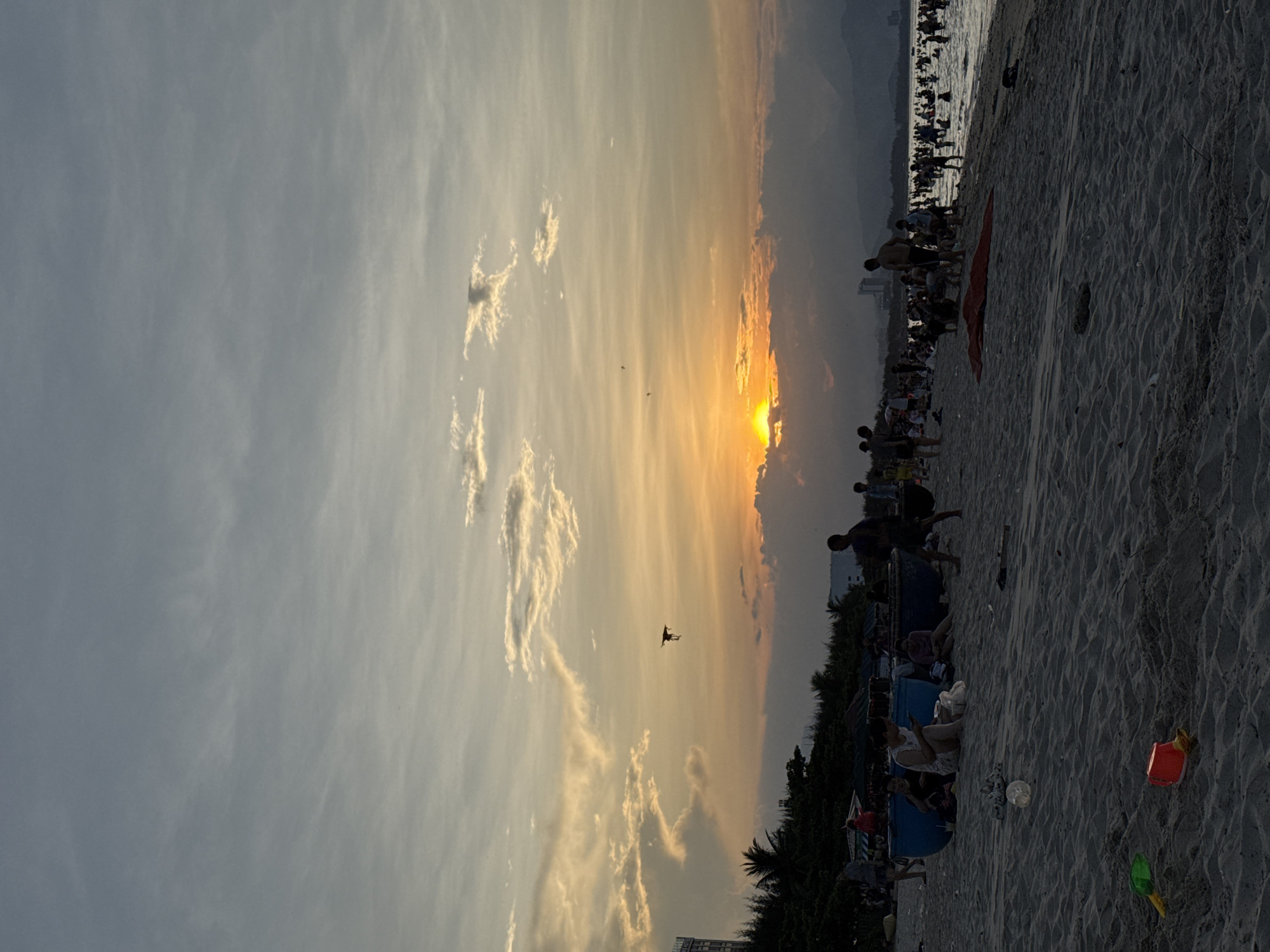 This image depicts a beach scene during sunset in Đà Nẵng, Vietnam. The sky is filled with a gradient of colors, from pale blue at the top to warm oranges and yellows near the horizon, with scattered clouds adding texture. The sun is low, partially obscured by clouds, casting a soft glow over the scene.
In the foreground, a sandy beach is visible, with ripples and textures created by the sand. Scattered on the beach are children's toys, including a green shovel and a red bucket. Further back, a group of people are gathered near blue boat-like structures, some sitting and interacting. Other individuals are walking on the sand or standing near the water's edge.
The middle ground of the image shows a multitude of people in the ocean, swimming and enjoying the water. The water appears calm with gentle waves. Along the coastline, a line of trees and some buildings are silhouetted against the sky. In the distance, mountains are visible, adding to the landscape. A kite is flying in the sky, adding a dynamic element to the scene. The overall atmosphere is one of leisure and relaxation, with people enjoying a beautiful sunset at the beach.