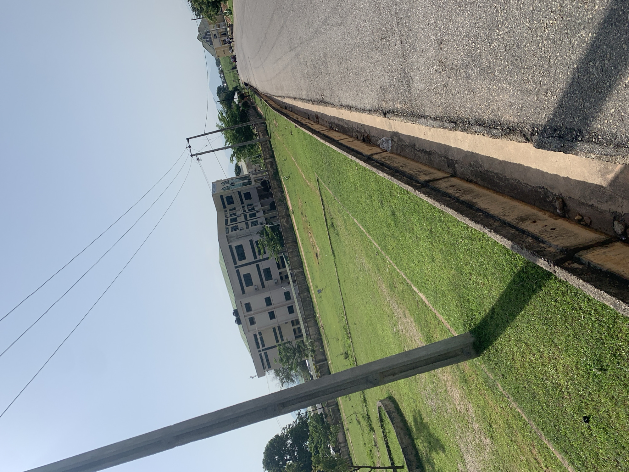 This image depicts a street scene in Jalingo, Nigeria, on a clear, sunny day. A paved road runs along the right side of the frame, bordered by a concrete drainage ditch. To the left of the road is a well-maintained grassy area, with a concrete utility pole in the foreground casting a long shadow. In the background, several buildings are visible, including a multi-story beige building with dark windows and a green roof, and a lighter-colored building with a prominent gabled roof and arched windows. The sky is a pale blue. There are a few cars and people visible in the distance near the buildings and on the road. The overall impression is one of an urban or semi-urban environment during daylight hours.