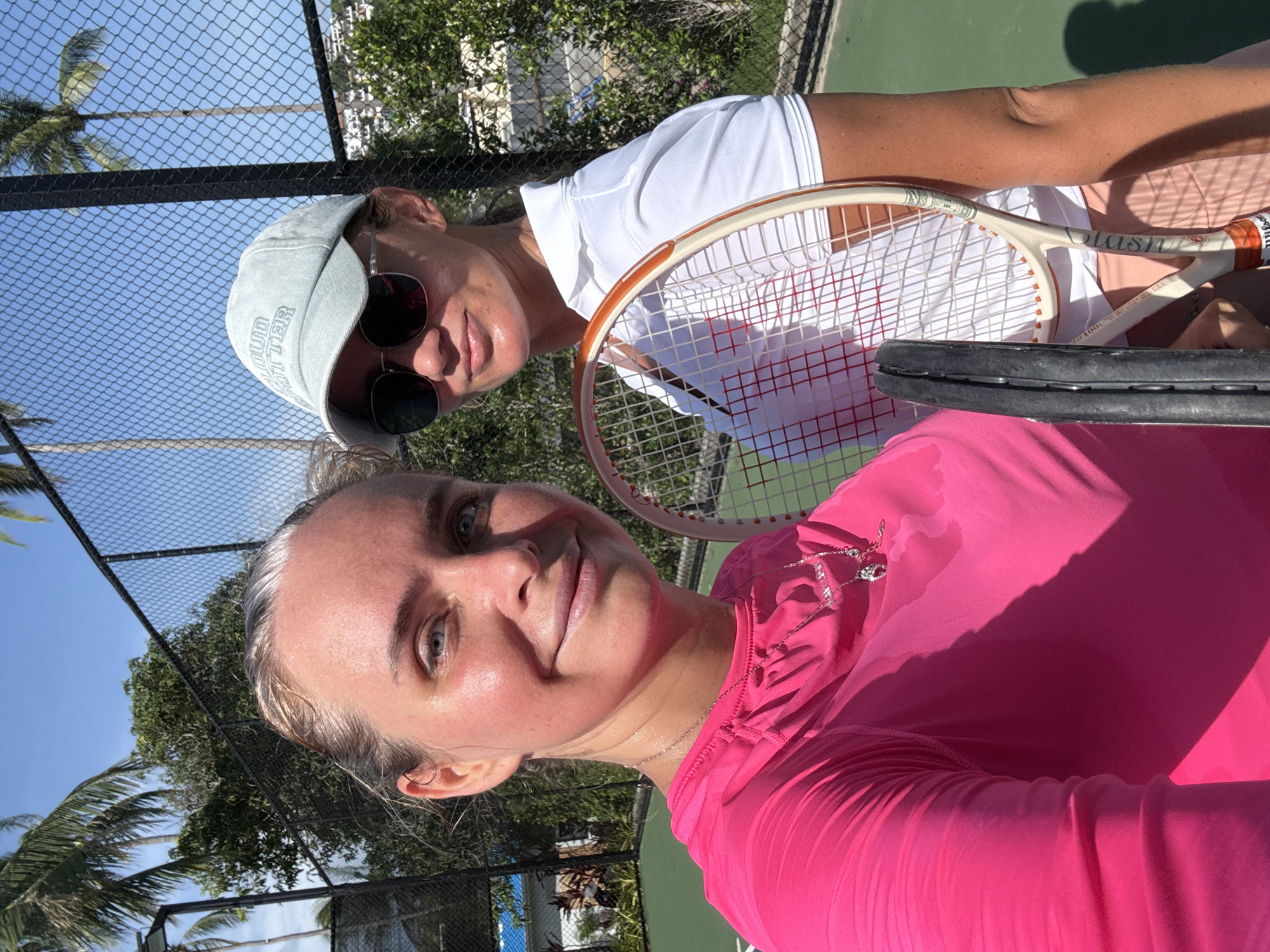 Two women are standing on a tennis court in Baan Plai Laem, Thailand. The woman in the foreground is wearing a bright pink long-sleeved shirt and a silver necklace. She has her hair pulled back and is smiling at the camera. The woman behind her is wearing a white polo shirt, a light blue baseball cap, and sunglasses. She is holding a tennis racket. The court is green and surrounded by a black fence. Palm trees and other greenery are visible in the background. The sky is blue and clear.