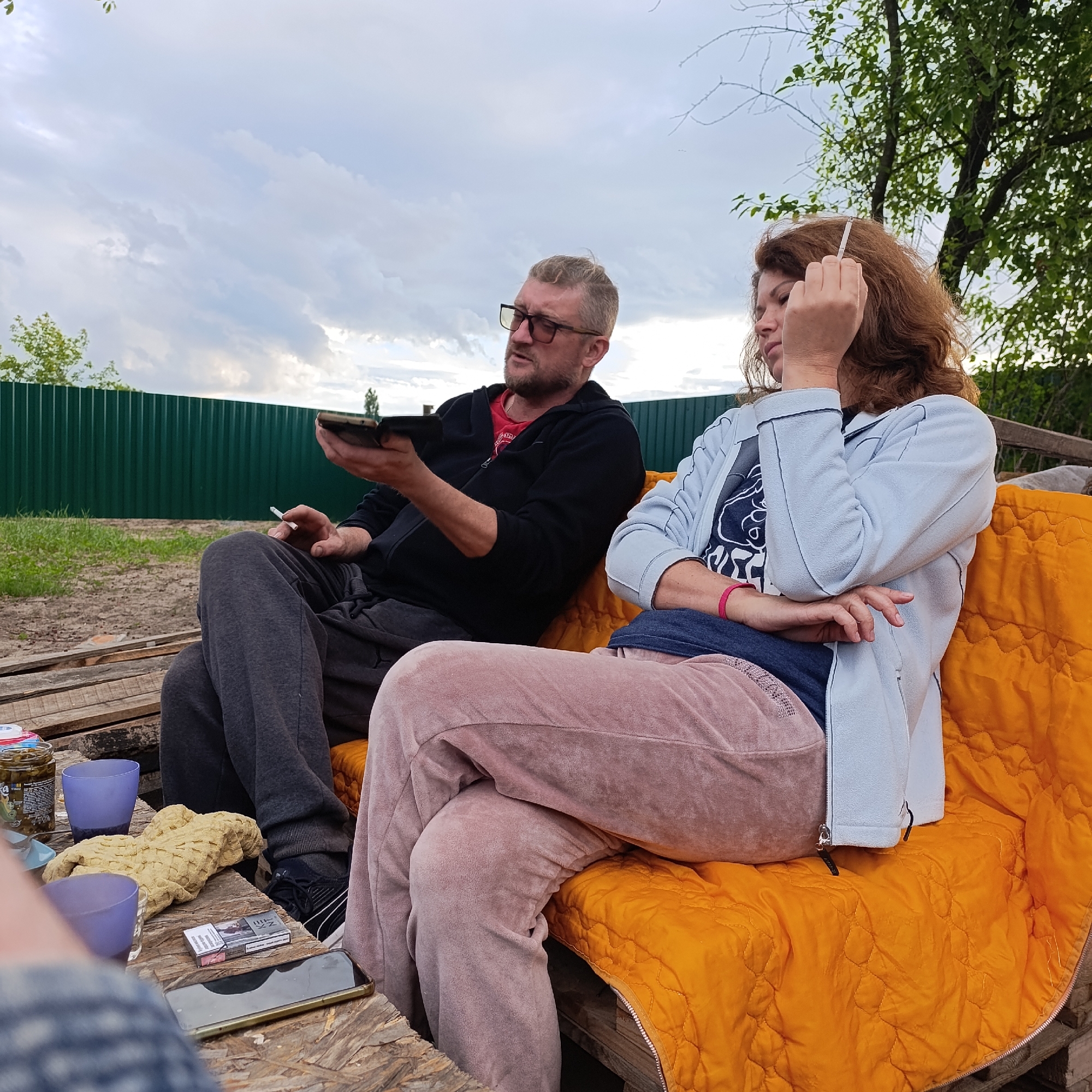 In Ladyzhyn, Ukraine, two adults are seated on a rustic, orange-upholstered couch, possibly constructed from pallets, outdoors. A man, wearing glasses and a black zip-up hoodie over a red t-shirt, sits to the left, holding a dark-colored mobile device. He has a cigarette in his right hand. To his right, a woman with reddish-brown, wavy hair is seated, holding a cigarette to her mouth with her right hand. She is wearing a light blue zip-up jacket over a dark t-shirt with a white graphic and pink sweatpants. A pack of cigarettes is visible on a wooden surface in the foreground, along with two purple cups and a jar. The background features a green corrugated metal fence and trees under a cloudy sky.