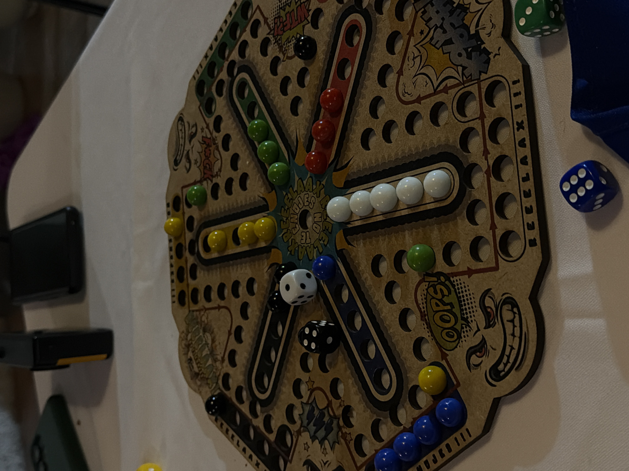 The image shows a close-up of a board game, likely Parcheesi or a similar game involving marbles, placed on a surface. The game board is a light brown color, with a hexagonal shape. The board has several circular indentations or holes, probably for holding marbles. There are marbles of various colors (white, red, green, yellow, and blue) placed on the board, in what appears to be designated starting or home positions. Dice, one blue and one green, are present near the edge of the board, along with additional dice on the board itself, implying active gameplay. The edges of the board have cartoonish graphics and phrases like "Oops!" and "REEEELAAAX!!!"
The setting appears to be indoors. The lighting seems soft and diffused. There are dark objects to the left of the game and blue cloth on the right, and they're located in a city of Unknown, Unknown.