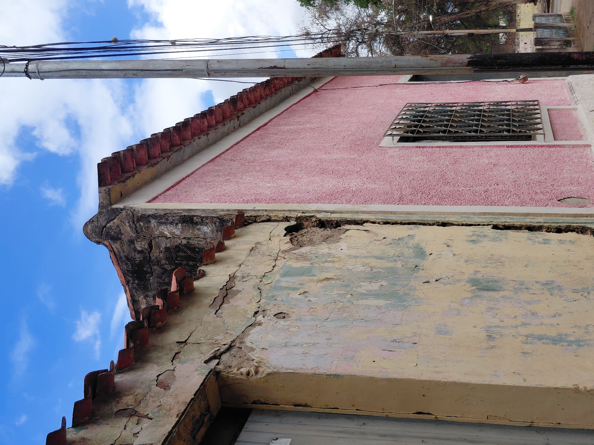 The image depicts a close-up, low-angle view of two adjoining buildings with distinct architectural styles and states of repair, under a partly cloudy blue sky. The building on the left, painted a faded yellow, shows significant signs of deterioration. Its exterior plaster is heavily cracked and peeling, revealing layers of older paint in pale green and pink hues. Parts of the wall are crumbling, exposing rough underlying material. A portion of a light-colored wooden door is visible on the left edge of this section. The roofline of this building is lined with red, weathered tiles, and the overhang shows exposed wooden beams and deteriorating plasterwork.
Adjacent to this is a building with a brightly painted pink exterior, appearing to be in better condition, though its stucco texture is visible. This building has a cleaner roofline with similar red tiles and a white border. A window with a decorative wrought-iron grate is present on this pink wall. To the right of the buildings, a tall, grey utility pole stands vertically, supporting a tangle of black electrical wires. Beyond the buildings and the utility pole, trees with bare branches are visible, suggesting a possibly dry climate or season. In the background, a low wall with decorative tiling can be seen, and a paved surface, likely a road or sidewalk, runs along the base of the buildings. The overall scene suggests an urban or semi-urban environment, possibly in a region with a history of older construction. The specific location in Santa Ana, Venezuela, is not visually evident from the image alone but is provided as contextual information.