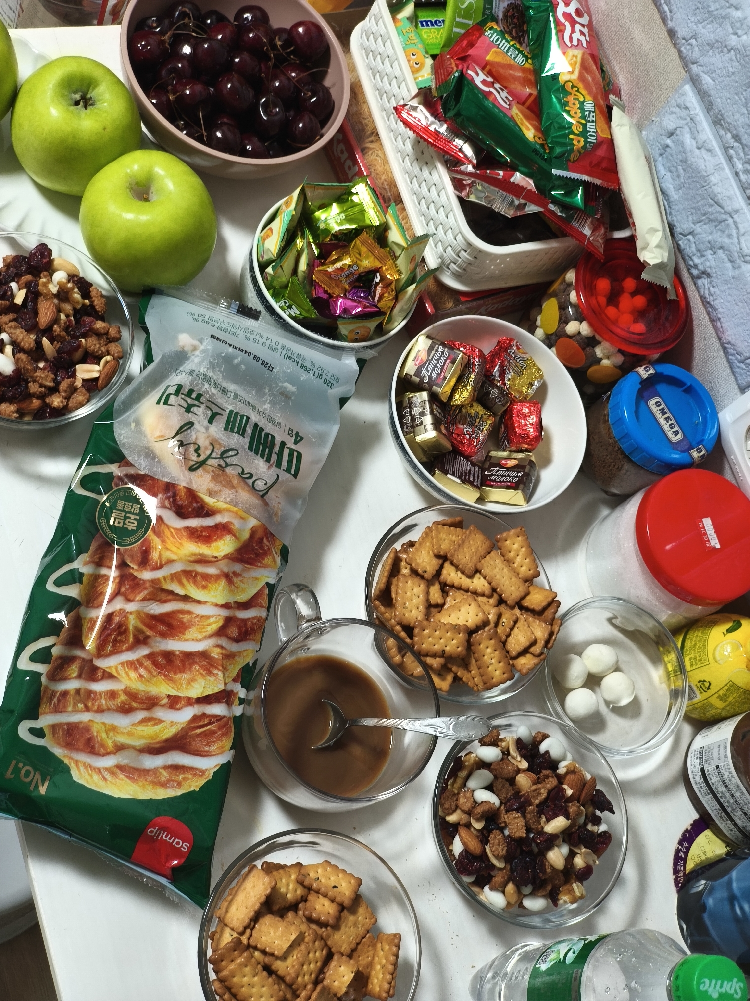 The image depicts a table laden with a variety of snacks and drinks. In the foreground, a large green bag with the text "No.1 samlip" and an image of pastries is prominent. To the left, two green apples sit in a white bowl. A bowl of cherries is positioned in the upper left quadrant. Several bowls contain an assortment of nuts and dried fruit, crackers, and small white balls that appear to be mozzarella balls. Various wrapped candies and snacks are scattered throughout the scene, some in a white basket. A glass cup contains a brown beverage, likely coffee, with a spoon. Two large plastic containers, one red and one blue, are visible in the background. A bottle of Sprite is also present. The overall setting suggests a casual gathering or a collection of snacks for consumption. There is no specific information to indicate the location is in Asan-si, South Korea.