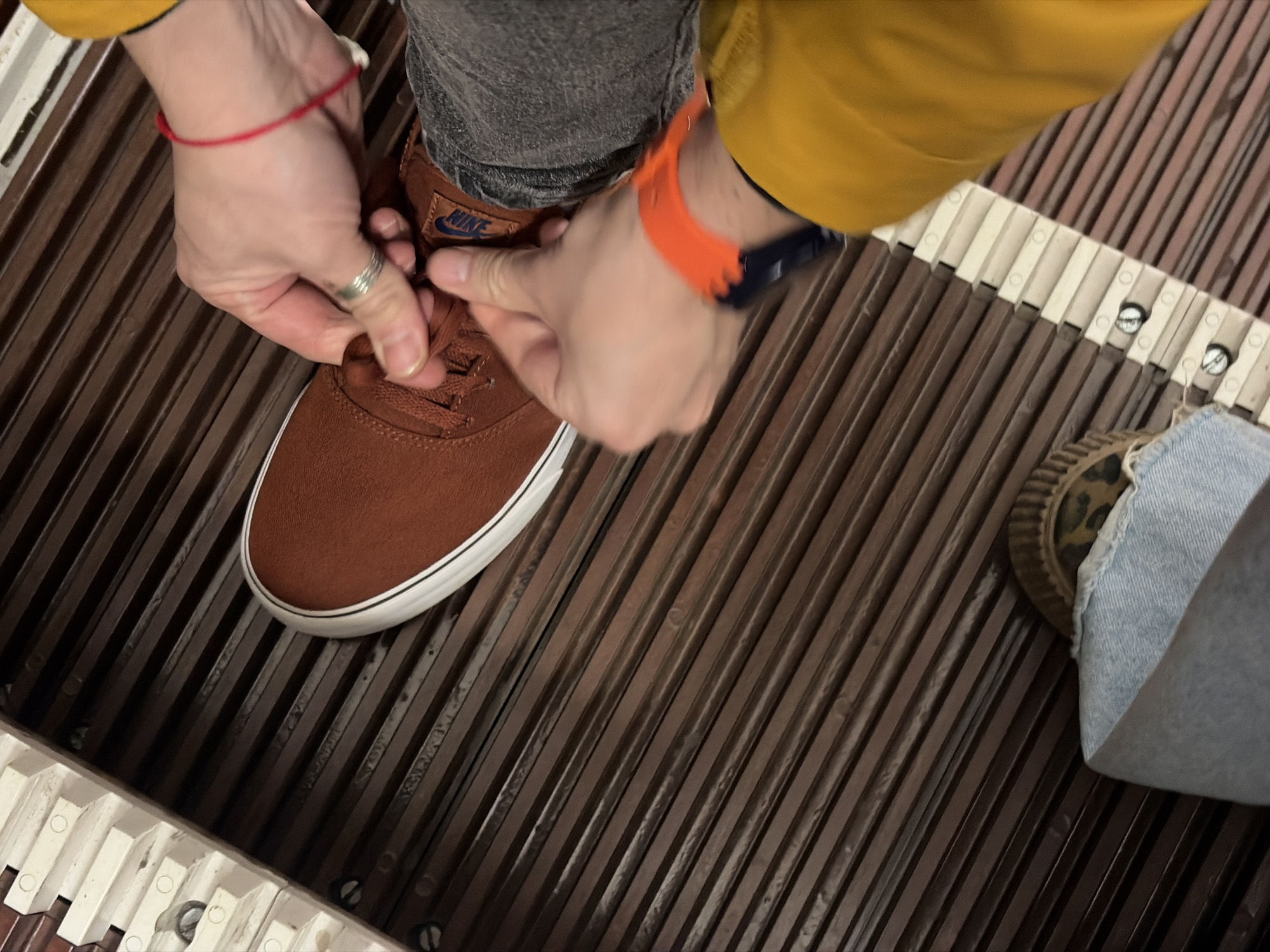 The image shows a person tying the laces of a brown Nike shoe while standing on an escalator. The escalator has a grooved brown metal surface with white horizontal borders. The person is wearing a yellow jacket, gray pants, and an orange wristband. A second shoe, partially visible, is a patterned sneaker with denim fabric covering part of it. The location appears to be inside an escalator in an indoor environment, though specific details to confirm a location in Saint Petersburg, Russia, are not present.