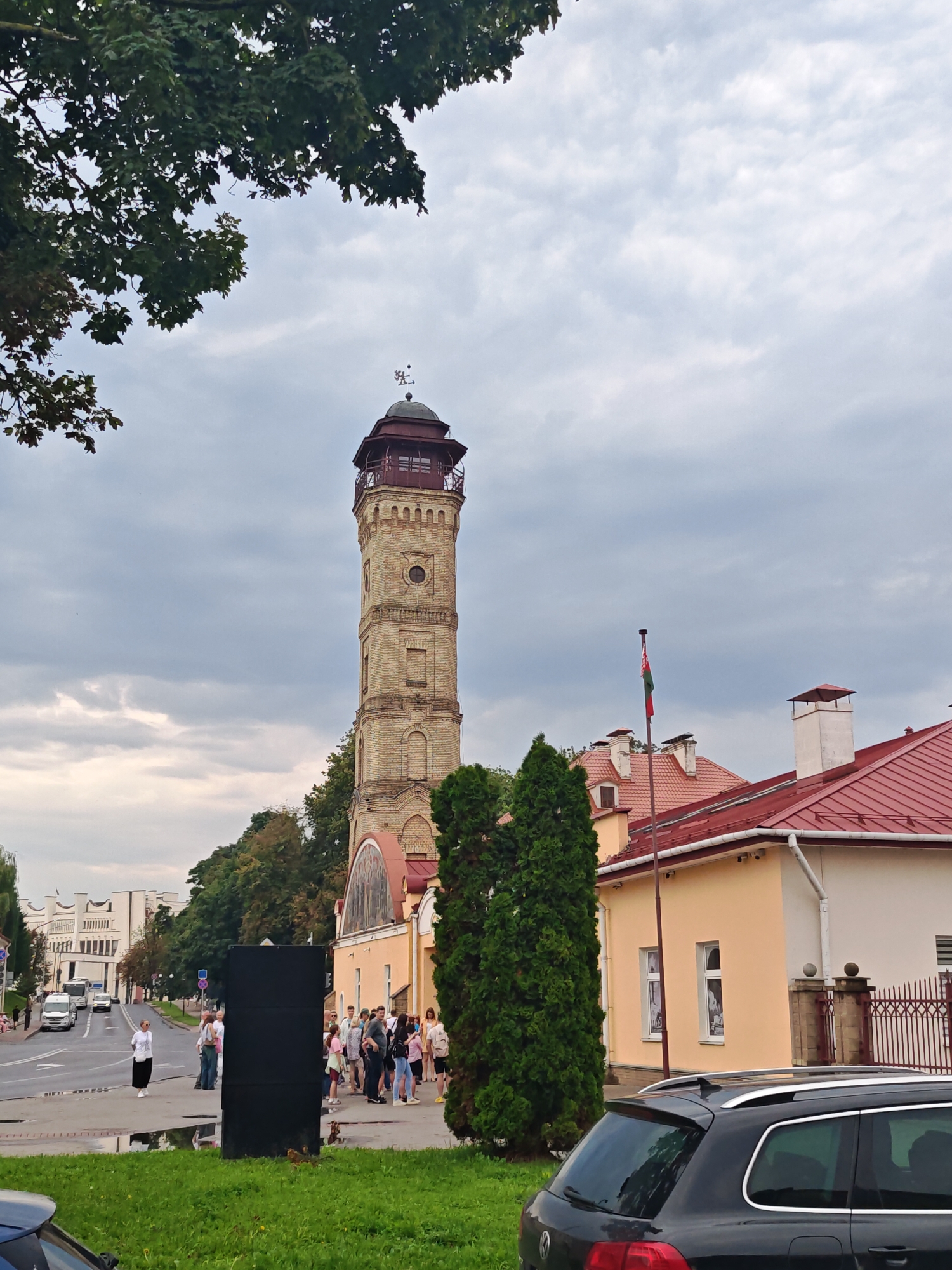 This image shows the Fire Tower in Hrodna, Belarus, on a cloudy day. The tower, made of yellow brick, has a dark red roof and is topped with a weather vane. To the right of the tower, a yellow building with a red roof is visible, featuring a flagpole with the Belarusian flag. In the foreground, a black SUV is parked, partially obscuring a grassy area. A group of people is gathered on the sidewalk near the yellow building, facing the street. On the street, several vehicles, including a bus and cars, are moving in the distance. The overall scene depicts an urban environment with historical architecture.
