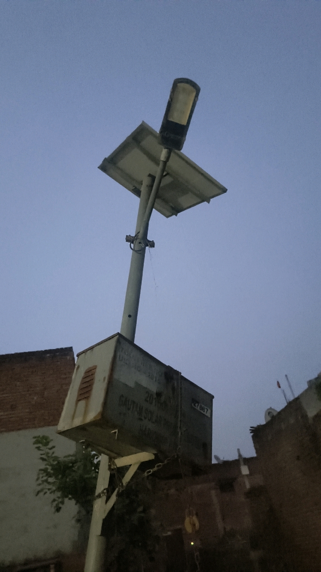 A low-angle, outdoor, vertical, full shot captures a solar-powered street light at dusk or dawn against a muted blue sky. The street light consists of a large, rectangular solar panel angled above a tall, grey pole. Attached to the pole, just below the panel, is a grey electrical box. Text is visible on the side of the box, including "2019 GAULTAM SOLAR POWER HARKIMPUR" and a number "27007". A chain is attached to the side of the box, extending downwards. To the left of the pole, a brick building is partially visible, and to the right, another building structure with a small flag is seen. The scene appears to be in an urban or semi-urban environment, possibly in Loni, India.