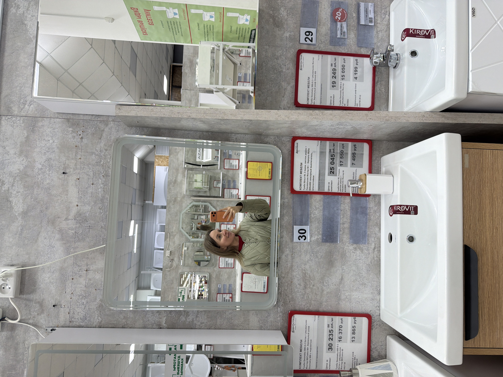 This image captures an indoor scene within a bathroom showroom or home improvement store, featuring several mirror and sink displays. The photograph is oriented vertically, rotated 90 degrees clockwise.
In the center, a woman is visible in the reflection of a large, rectangular mirror with rounded corners, holding an orange-cased smartphone and appearing to take a selfie. She is wearing a light olive-green quilted jacket over a red top and has light brown hair. Her reflection shows her smiling slightly as she poses. The mirror itself is part of a display set, reflecting the store's ceiling and other display mirrors in the background.
Below this mirror, a white rectangular sink with a chrome faucet is mounted on a wooden-toned cabinet. Adjacent to it, a red price tag labeled "30" provides details in Russian, including "КОМПЛЕКТ МЕБЕЛИ" (Furniture Set) and lists items like "Тумба Кёльн 60," "Умывальник Фостер 60," and "Зеркало Malta 600х600 сенсорный выключатель, влагостойкое" with prices of "25 045 руб./шт" for the set, "17 550 руб," and "7 495 руб." The brand "KIROVIT" is visible on the sink.
To the left of this display, a white electrical outlet and wires are visible on the grey, textured wall. Above the central display, another similar bathroom set is shown, with a white sink and a mirror reflecting the store interior. Its red price tag, labeled "29," specifies "Тумба Geometry 500-0-2 + умывальник Фостер 50+ Зеркальный шкаф Grand 450," with a set price of "19 249 руб./шт." A "-20%" discount tag is also attached. Another display is partially visible at the bottom.
Visible text on various signs includes store or brand names like "ДОБРОСТРОЙ," "AZAR," and "VIGO," along with product specifications and prices in Russian rubles, confirming the location in Chaikovsky, Russia.