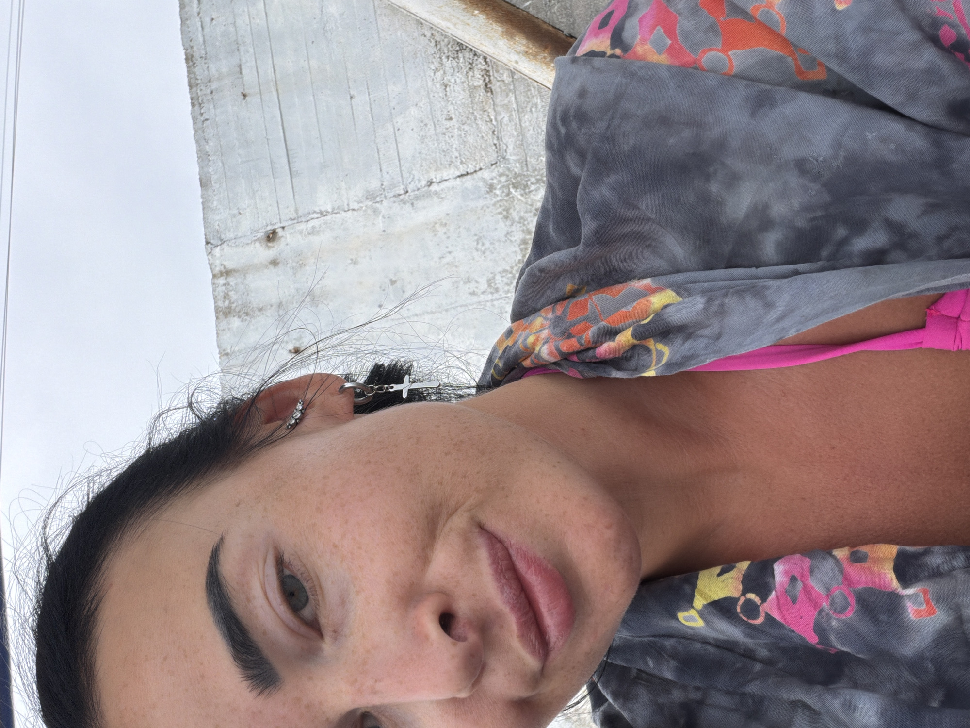 A close-up, angled shot shows a woman with dark hair and fair skin, adorned with freckles. She is wearing a gray, tie-dye robe with a colorful, abstract pattern in shades of pink, orange, and yellow, revealing a bright pink bikini top underneath. Her left ear features multiple piercings, including a small, silver ear cuff with three clear stones and a hoop earring with a dangling silver cross. She is looking towards the camera with a subtle smile, her head tilted slightly. Behind her, a textured, light-colored concrete structure forms the background, with a rusted metal pipe visible on the right side of the frame. The sky above is overcast and pale gray. The overall impression is a casual, outdoor setting, possibly a resort or beach area, but without specific landmarks to confirm a location in Sochi, Russia.