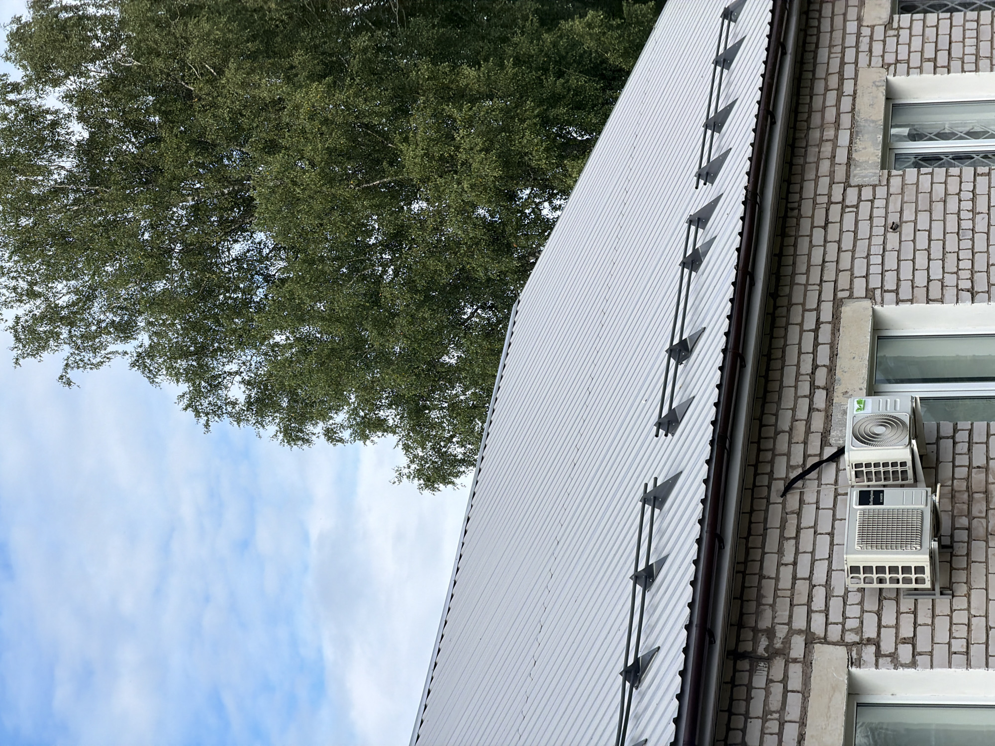 The image shows the side of a brick building with two air conditioning units mounted on the wall. The building has a corrugated metal roof with a series of triangular brackets attached to it. A large tree with green leaves is visible behind the roof, and the sky is blue with some white clouds. The scene appears to be outdoors during the daytime. The perspective is from a low angle, looking up towards the building and the tree. There is no visible text in the image. The context provided is Moshenskoe, Russia.