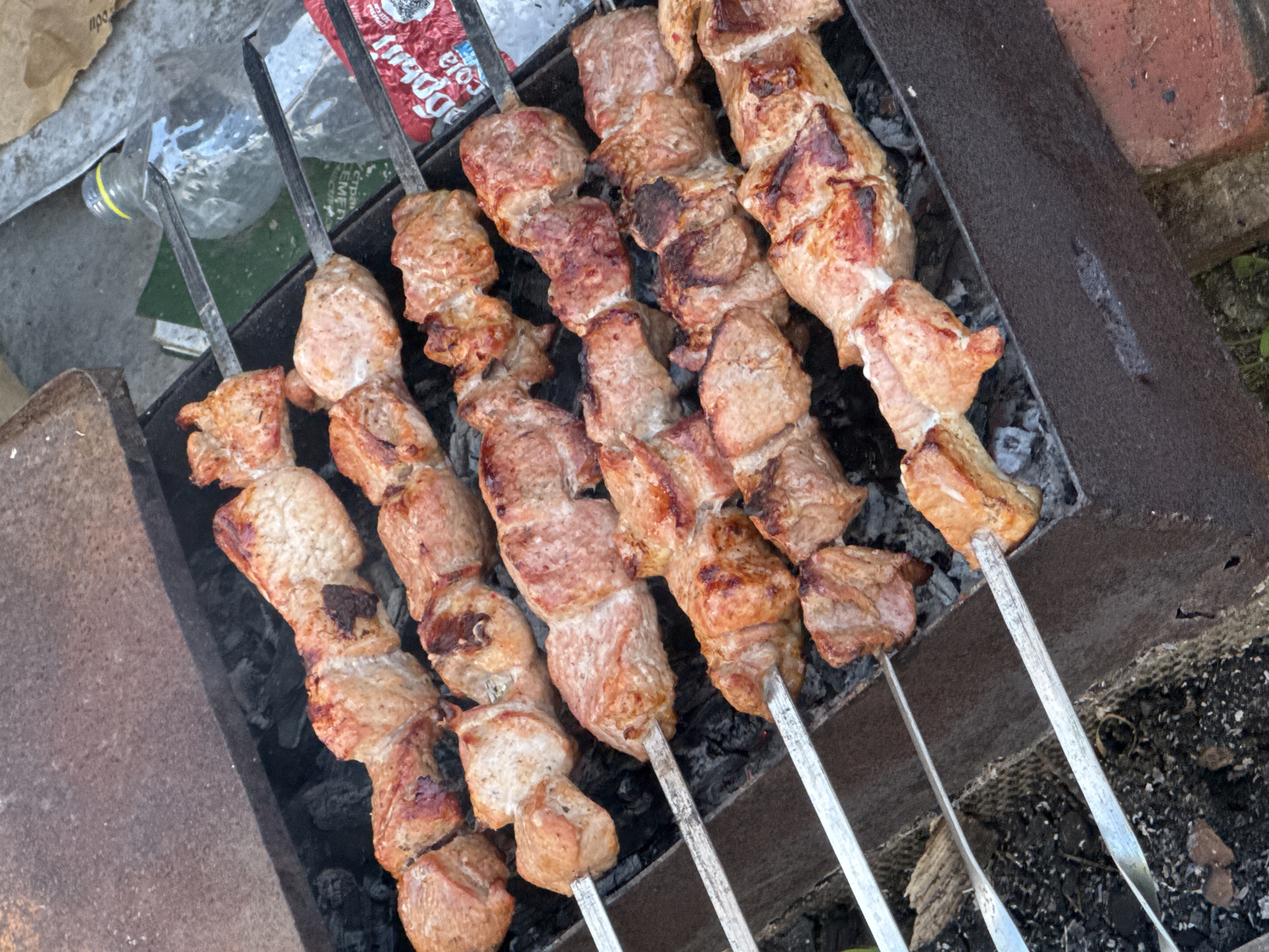 This is a close-up, overhead shot of meat skewers cooking over charcoal in a metal brazier. The skewers are arranged horizontally and fill most of the frame. The meat appears to be pork, cut into cubes, and is browned and slightly charred in places. The brazier is dark, rectangular, and shows signs of use. Scattered charcoal is visible beneath the skewers, some glowing red. In the background, to the right, a clear plastic bottle and a torn red and white wrapper with Cyrillic text are partially visible. The overall scene suggests an outdoor grilling activity. There are no people visible in the image. The location is identified as Bigila, Russia.