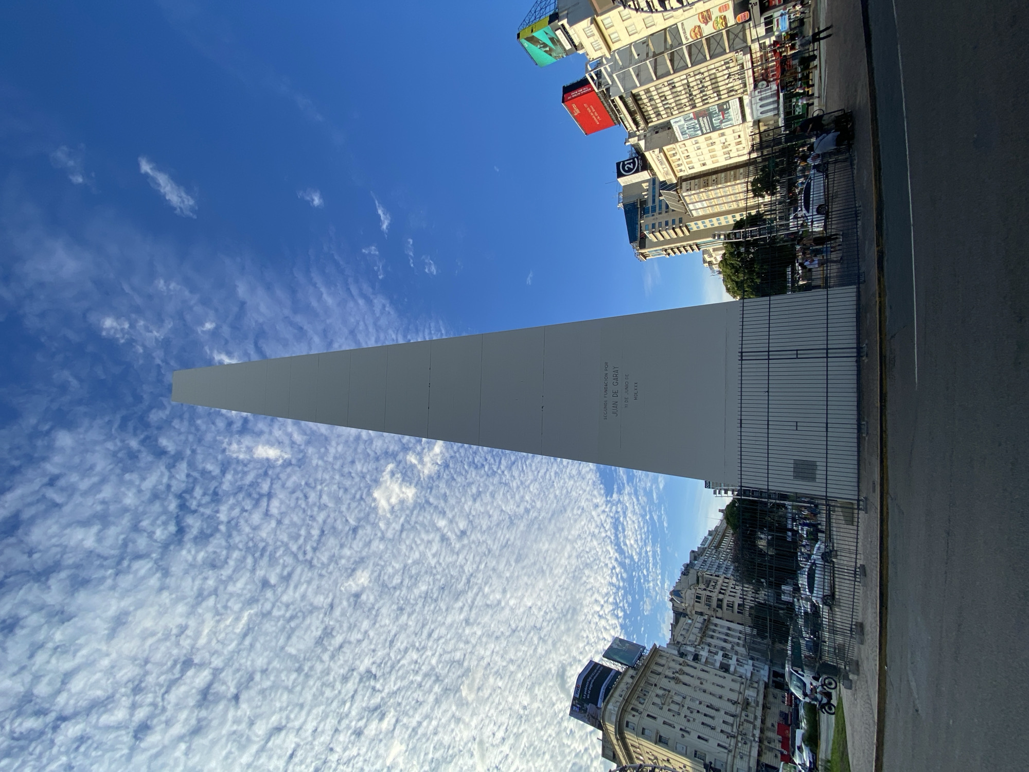 The image shows the Obelisco de Buenos Aires, a national historical monument in the city. The obelisk is white, rising against a blue sky with scattered clouds. Adjacent to the obelisk are tall buildings with commercial signage and advertisements. There are people present, though they are small relative to the scale of the obelisk and buildings. The environment is an urban setting with a paved street.