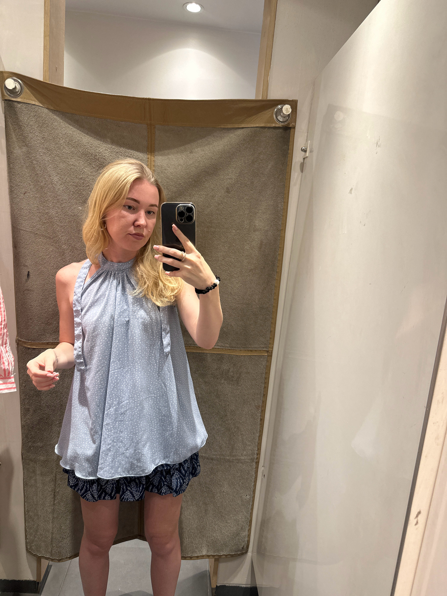A young woman with blonde hair stands in a fitting room, taking a mirror selfie with her phone. She is wearing a light blue, sleeveless, polka-dotted halter top and dark blue patterned shorts. The fitting room has a brown fabric backdrop and a white wall.