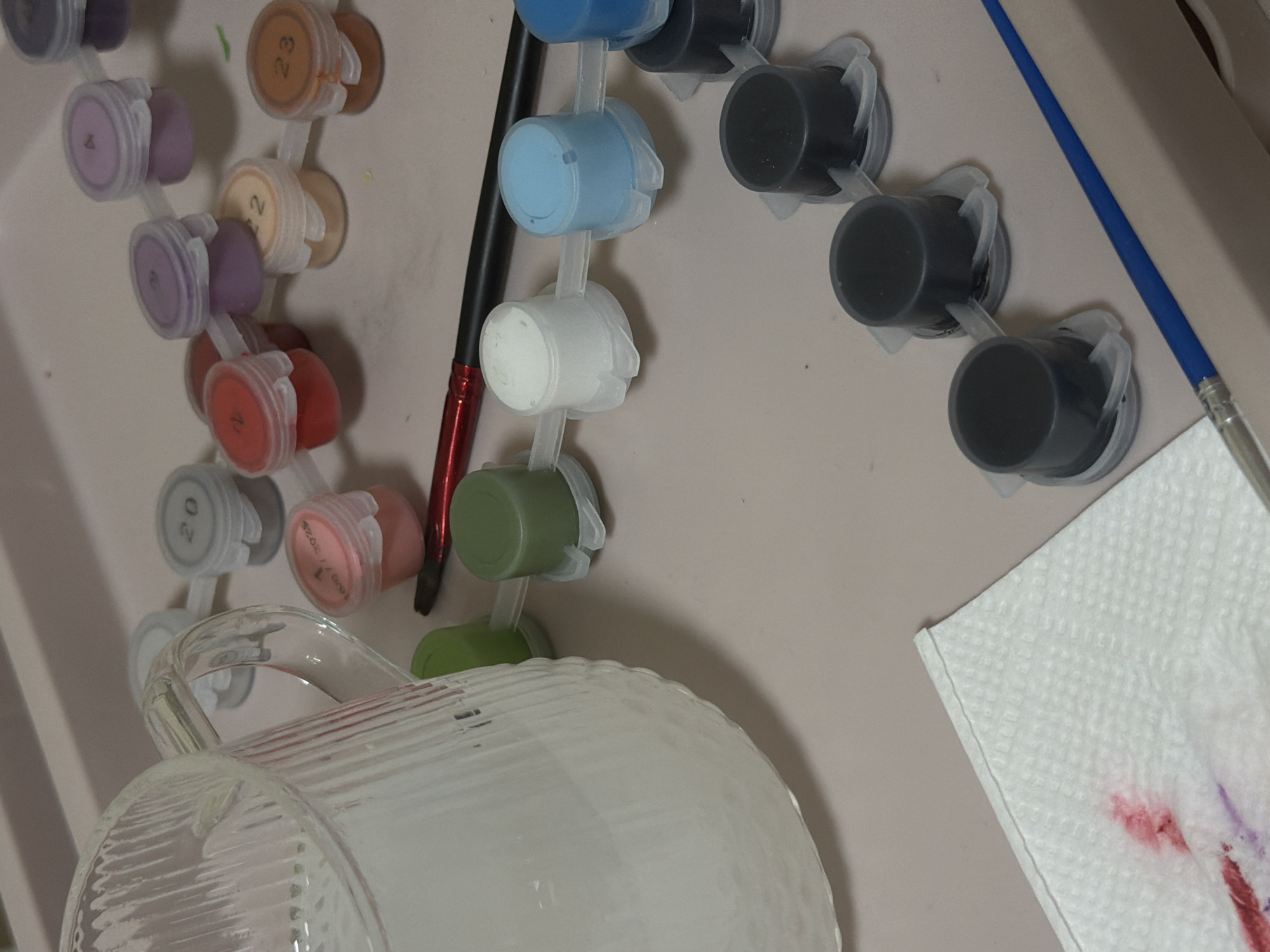 The image shows a paint-by-numbers setup on a light beige surface. A container filled with water is in the lower left. Aligned across the top are paint pots in a connected series, with colors ranging from purples and oranges on the left, to blues, whites, greens, and blacks on the right. A red and black paintbrush and a blue-handled paintbrush are also visible. A white paper towel with red and purple stains is in the lower right. There is no indication of Izhevsk, Russia.