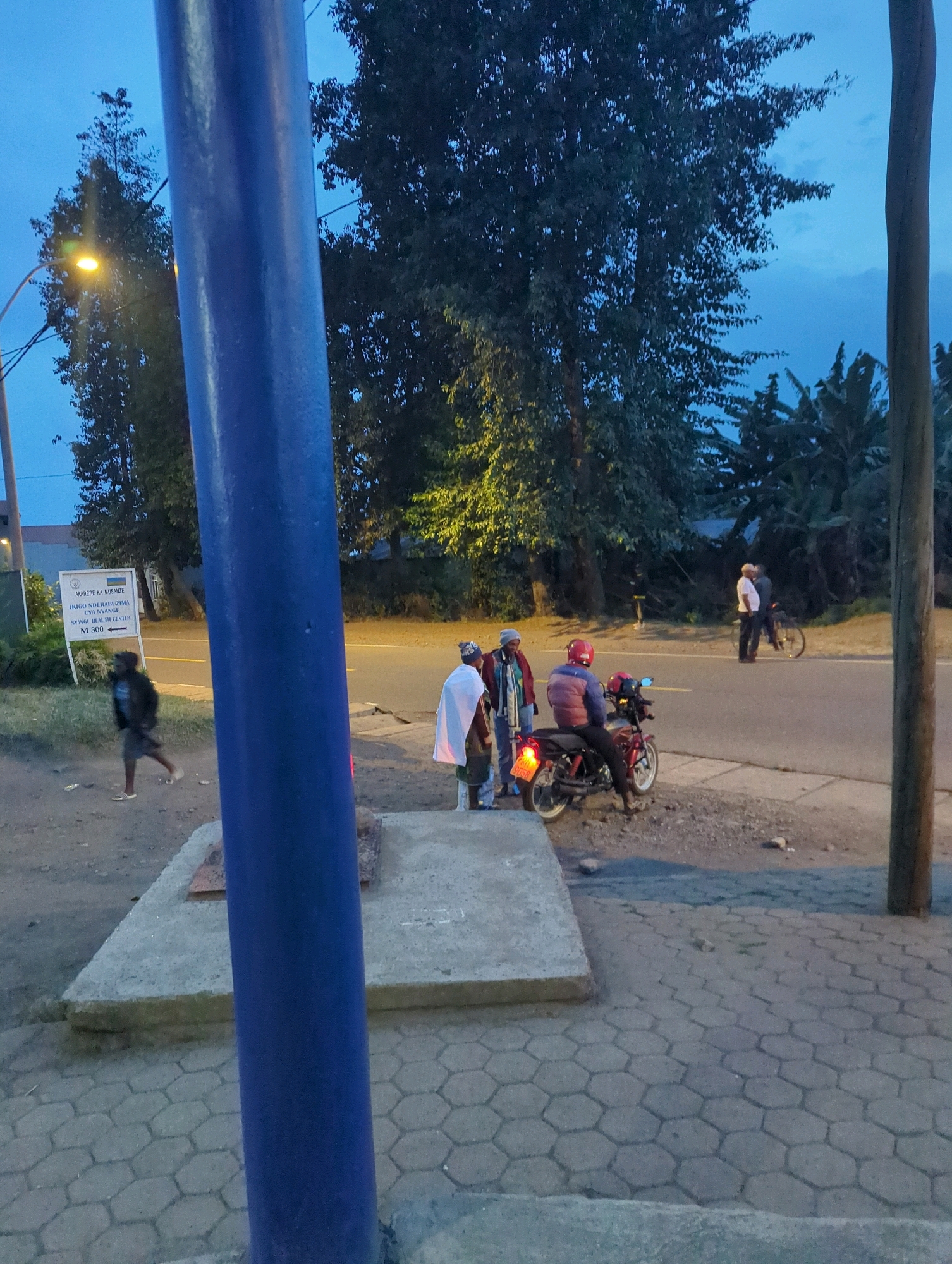 The image shows a scene in the city of Musanze District, Rwanda, with a blue pole in the foreground. On the side of the road, a group of people are gathered. One person is riding a motorcycle, while others stand nearby. A sign is visible on the left, indicating "AKAREBE KA MUSANZE IKIGO NDETSE UZUIMA CY'INYAYU NYAMYASA HEALTH CENTER M 300" and an arrow pointing right, suggesting the location of a health center. The environment includes trees, a road, and a sidewalk with hexagonal paving stones. The lighting suggests it is dusk or early morning.