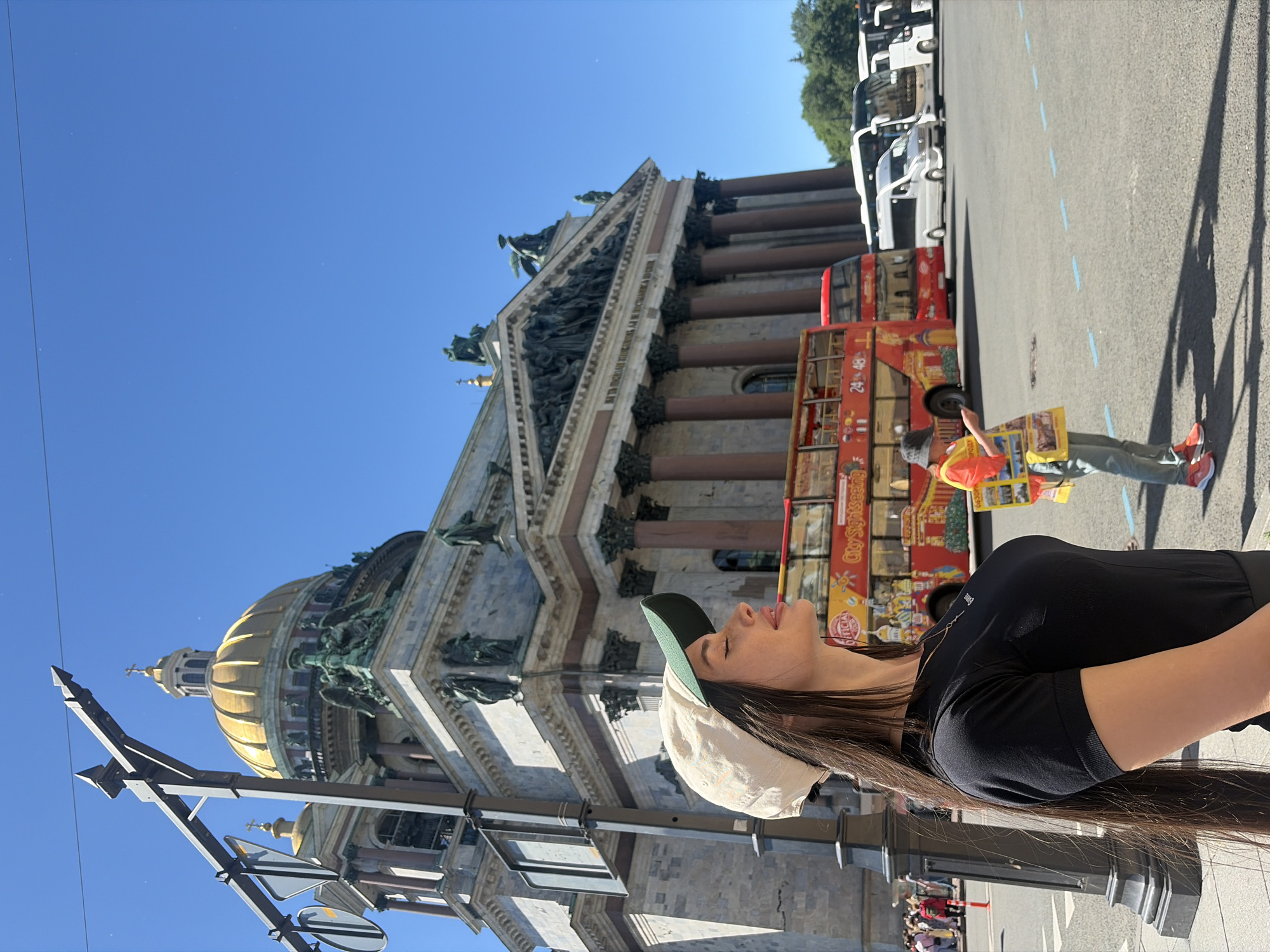 In Saint Petersburg, Russia, a woman wearing a black short-sleeved shirt and a beige baseball cap with a green brim stands in the foreground. She has long dark hair and her eyes are closed, with her face tilted upwards. Behind her, a large building with a golden dome and classical architecture is visible, likely Saint Isaac's Cathedral. A bright red double-decker tour bus with "City Sightseeing" text and several white vans are parked in the square. A tour guide wearing an orange vest is standing to the right, holding a pamphlet. The scene is set on a clear, sunny day.