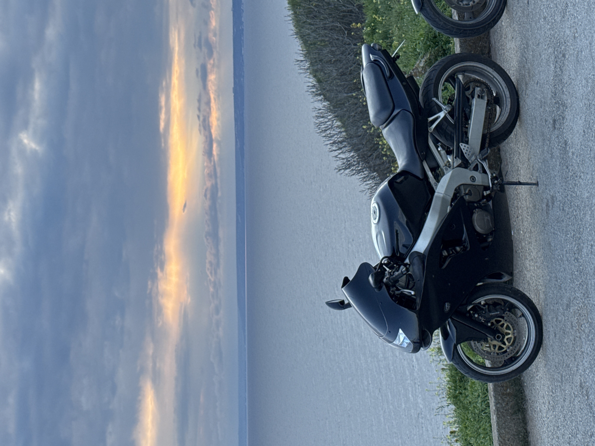 The image shows two motorcycles parked on a paved road or path near a body of water. The background features a sky with clouds and a strip of sunlight on the horizon. The landscape appears to be flat. The presence of yellow flowers along the roadside suggests a natural environment. The location is possibly near Balgarevo, Bulgaria.