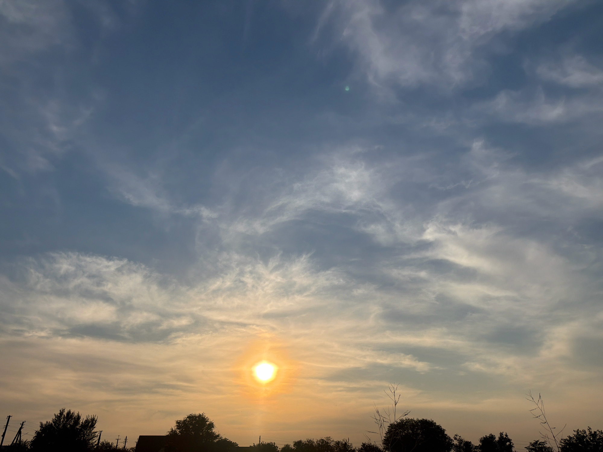The image shows a sky with a sunset. The sun is visible in the lower center of the image, casting a golden light. The sky is filled with clouds, some of which are wispy and thin, while others are darker and more dense. In the foreground, silhouetted against the bright sky, are trees and some structures that appear to be houses or buildings. The overall impression is one of a peaceful evening scene. The image does not provide any specific details about the location within the city of Unknown, Unknown.
