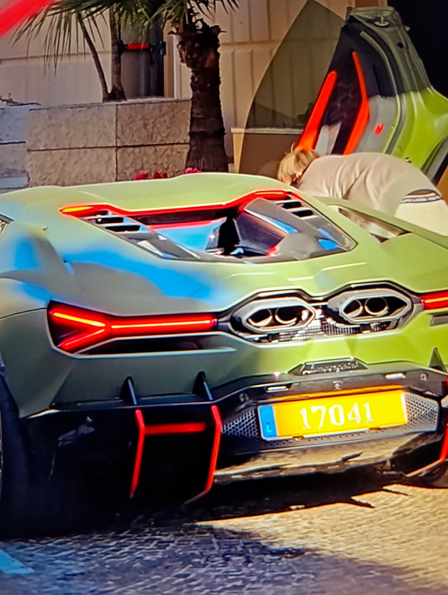 The image shows the rear of a green Lamborghini Sian with a Monaco license plate reading "17041." The car is parked on a cobblestone street. A person is bending down near the open driver's side door. The background includes foliage and a building facade.