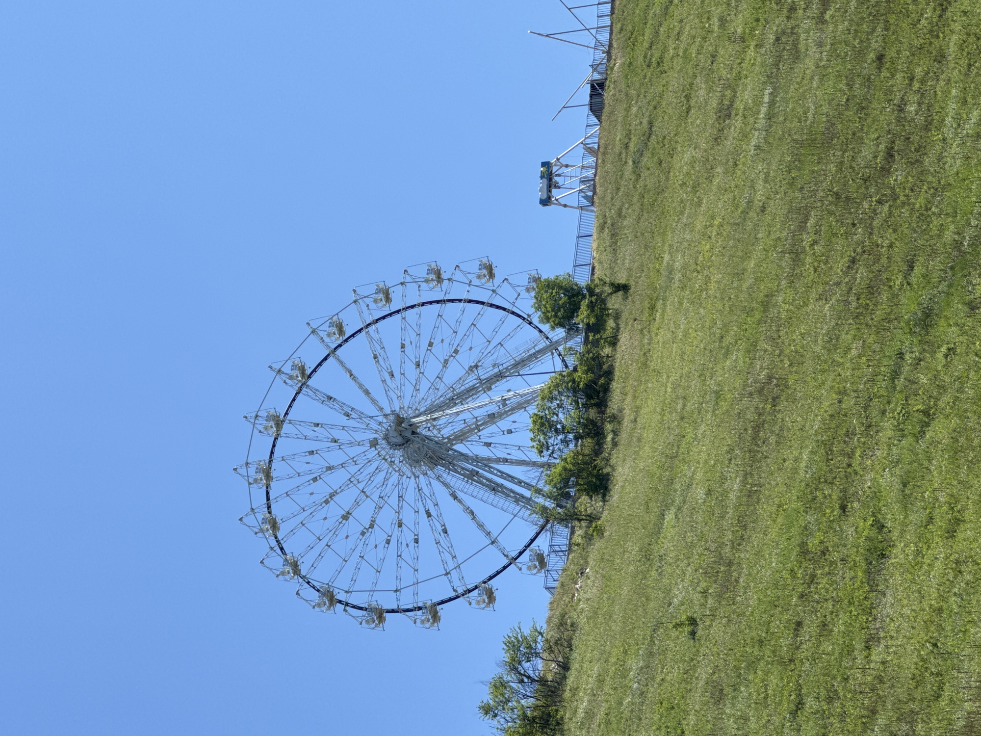 A large Ferris wheel stands prominently against a clear, brilliant blue sky, situated in Gelendzhik, Russia. The Ferris wheel, a complex structure of white metal spokes and supports, occupies the left side of the frame. Numerous passenger gondolas are suspended around its rim, appearing empty.
To the right, a verdant green grassy hill or slope rises, creating a natural backdrop for the mechanical attraction. The grass shows varying textures and shades of green, suggesting a well-maintained landscape. Several small trees and bushes are scattered near the base of the Ferris wheel and along the edge of the grassy area, adding natural elements to the scene. Parts of a light-colored structural platform or railing are visible atop the hill, hinting at further infrastructure or access points.
The bright, cloudless sky indicates a sunny day with excellent visibility, likely morning or early afternoon given the even lighting. There are no people visible in the image, nor is any discernible text present. The scene conveys a sense of pleasant weather and an active outdoor leisure environment, typical of a tourist attraction or amusement park.