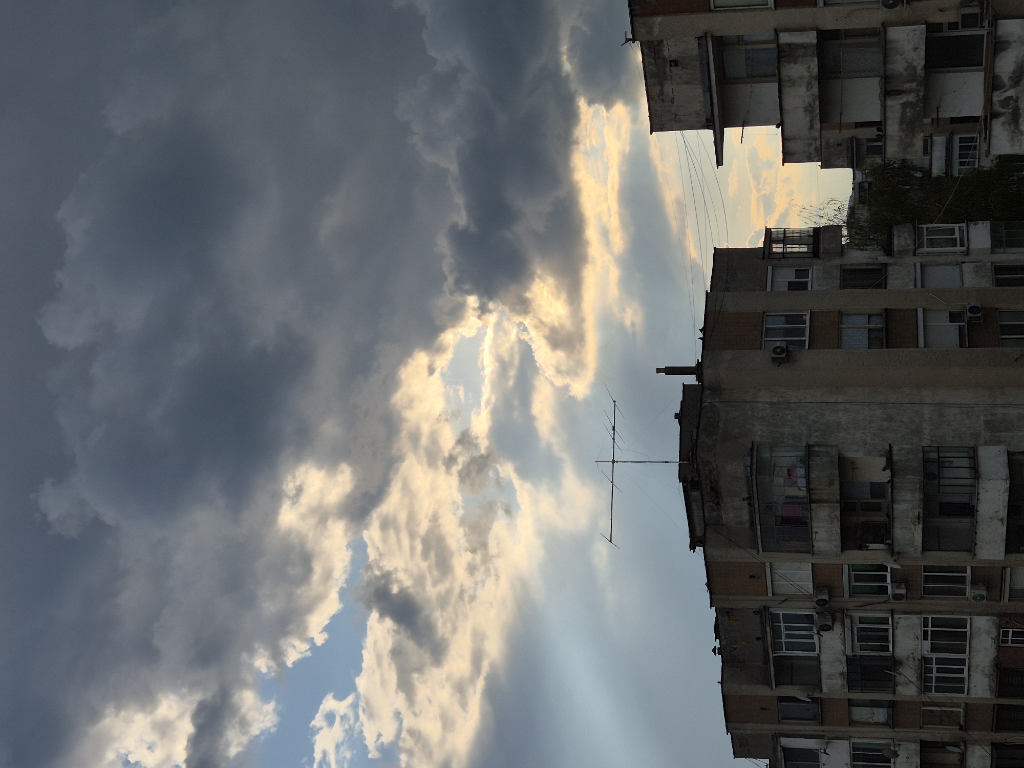 The image displays a dramatic sky over residential buildings, likely in an urban setting in Pavlikeni, Bulgaria. The sky is the dominant feature, filled with a striking contrast of dark, heavy storm clouds on one side and brilliant, golden-white clouds on the other, illuminated by sunlight breaking through. Visible rays of light pierce the cloud cover, and patches of blue sky can be seen.
Below the dynamic sky are two multi-story buildings, typical of utilitarian, possibly post-WWII or communist-era architecture, characterized by concrete facades, numerous windows, and balconies. They show signs of age and wear. Several external air conditioning units are visible on the building exteriors. A large, multi-element antenna extends from one of the buildings into the sky, and thin wires crisscross the scene. Some sparse vegetation or ivy grows on one of the building walls.
The time of day appears to be late afternoon or early evening, suggested by the warm, golden light filtering through the clouds, indicative of a sunset or sunrise. The weather is highly dynamic, capturing a moment of transition between dark, brooding clouds and bright, sunlit skies, creating a powerful and atmospheric scene. There is no human activity or specific event taking place, and no visible text is present.