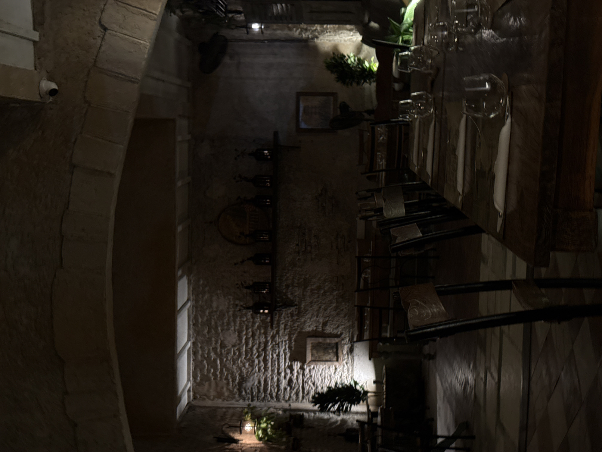 The image shows the interior of a restaurant in Unknown, Unknown. The walls are textured with a rough, stone-like finish. There is an arched entryway leading into the restaurant from the left, and a security camera is mounted near the arch. Inside, a dining table is set with glassware and linens. Shelves holding wine bottles are visible on the back wall, and a framed picture hangs above a small plant. The lighting is dim, creating a warm atmosphere. Tables and chairs are set up in the background, suggesting a functional dining area.