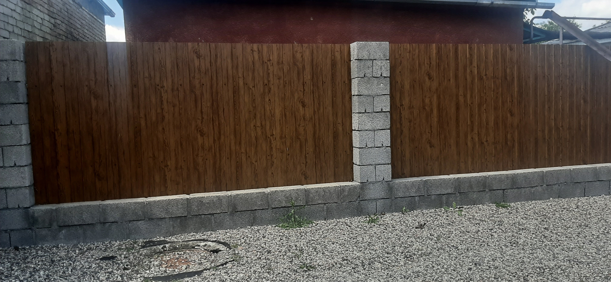 The image depicts a wooden fence with a wood grain pattern, erected on a concrete block foundation. The fence is supported by concrete block pillars, one of which is prominently visible in the center of the frame. The ground in front of the fence is covered in gravel, with a circular manhole cover visible on the left side. In the background, a red-colored building is partially visible, with a portion of a roofline and a gutter system. The sky above is partly cloudy. The scene appears to be an outdoor area, possibly a backyard or a property boundary, located in the city of Yessentukskaya, Russia. There are no people or animals visible in the image. The overall impression is one of a well-constructed, functional fence and foundation.