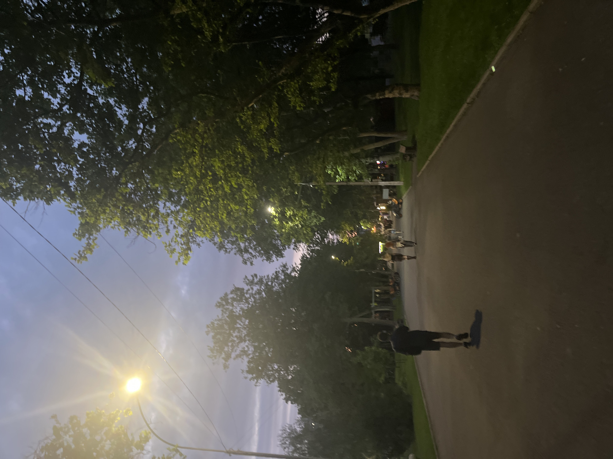 The image depicts a park at dusk, likely in Krasnodar, Russia, given the provided context. A paved pathway stretches into the distance, flanked by mature trees casting shadows. The sky is a dusky blue with streaks of pale clouds, and a bright streetlight on the left illuminates the scene, creating a lens flare.
In the foreground, a lone individual, silhouetted against the dim light, is walking away from the camera. They appear to be a male, wearing dark shorts and a dark t-shirt. His shadow is cast long on the pathway. Further down the path, a small group of people can be seen walking, their figures less distinct. To the right of the pathway, there is a grassy area with a park bench visible. In the background, more trees and hints of park structures or stalls suggest a public recreational area. The overall atmosphere is calm and suggestive of an evening stroll or exercise.