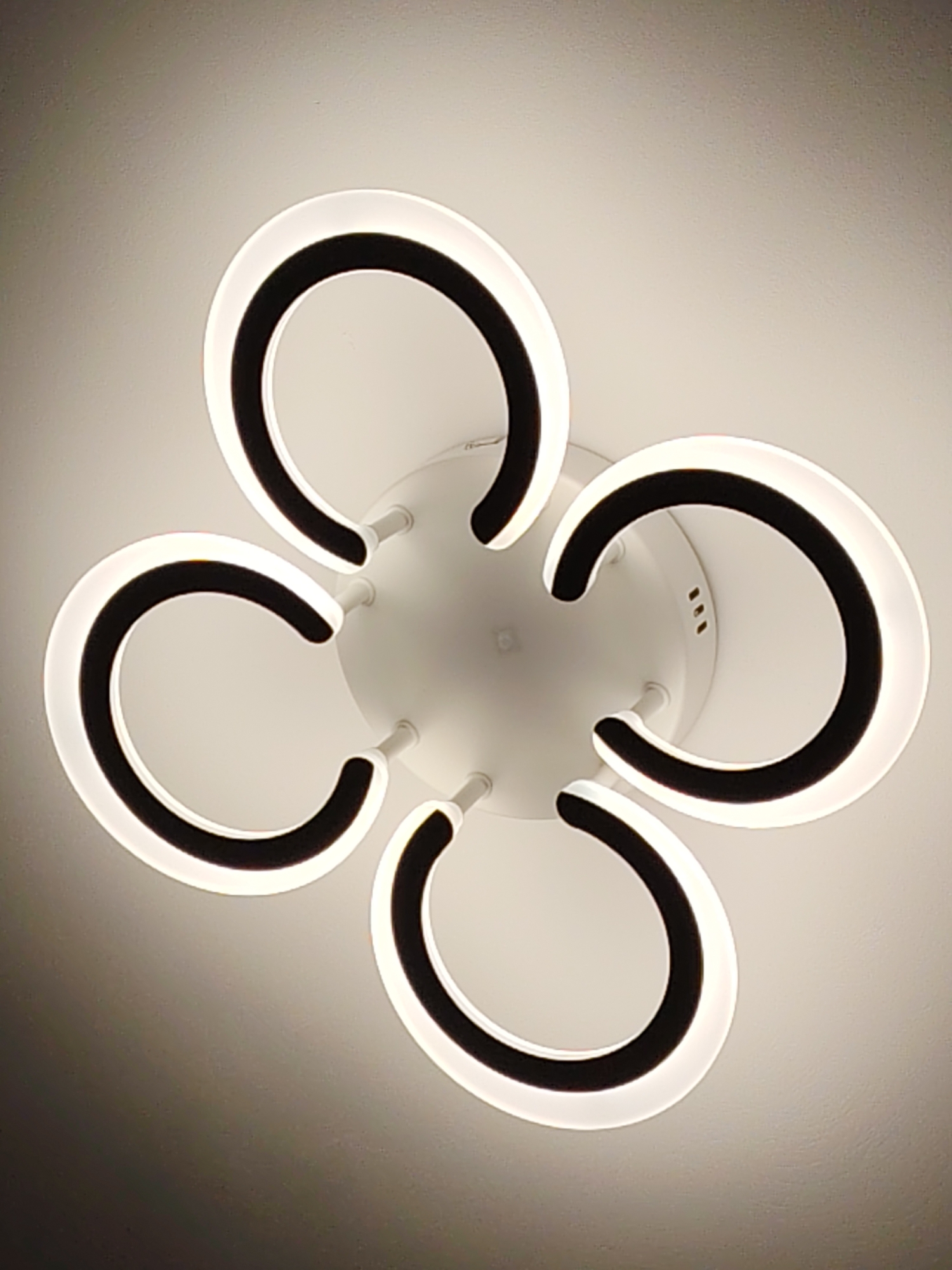 A modern, five-armed LED ceiling light fixture is centrally positioned against an off-white, textured ceiling. The fixture, located in Yekaterinburg, Russia, features a white circular central base from which five C-shaped elements radially extend. Each C-shaped element is connected to the central base by a slender white rod. The convex outer edge of each C-shaped element emits a bright white light, contrasting with its dark, opaque concave inner edge. The central base presents three small, elongated rectangular perforations. The fixture illuminates the surrounding ceiling with a diffused white light.