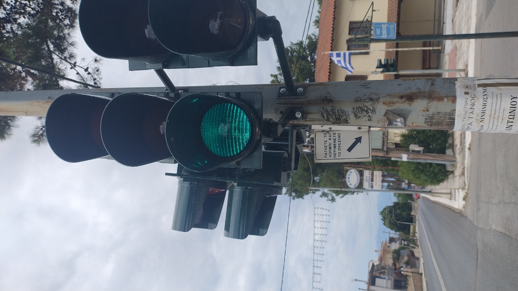 A traffic light showing a green signal is positioned on a metal pole in Νέα Κερασιά, Greece. The traffic light has multiple lenses, with the bottom lens illuminated in green. A white sign with black text and an arrow pointing left is attached to the pole below the traffic light. The sign reads "ΠΑΤΗΣΤΕ ΤΟ ΚΟΥΜΠΙ ΚΑΙ ΠΕΡΙΜΕΝΕΤΕ ΤΟ ΠΡΑΣΙΝΟ" which translates to "PRESS THE BUTTON AND WAIT FOR THE GREEN." In the background, there is a street, buildings with red-tiled roofs, trees, and a Greek flag. The sky is overcast.