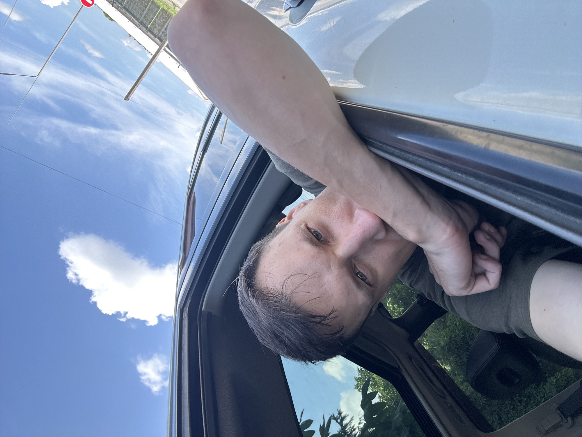 A man is leaning out of a car window, his arm resting on the door frame. He is looking at the camera with a thoughtful expression, his hand near his mouth. The car is parked outdoors, with a blue sky and white clouds visible above. Green trees and foliage can be seen through the car window. The overall impression is one of a candid moment captured on a sunny day. The location is Shatava, Ukraine.