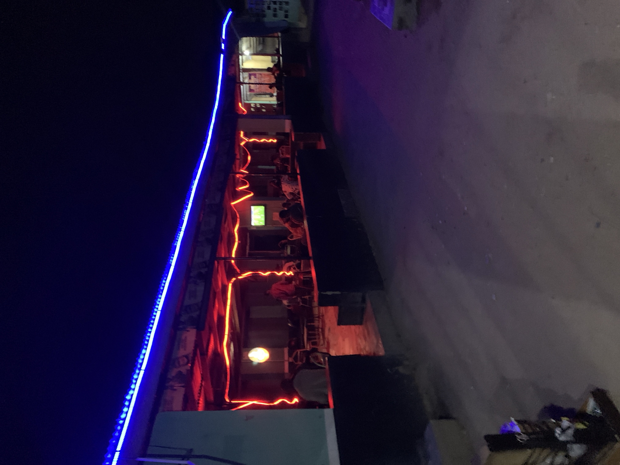 Here is a detailed description of the image:
**Scene:** An outdoor restaurant setting at night, possibly in the vicinity of New Stadium, Nigeria, given the location information.
**Environment:** The scene is dimly lit. A building with a facade decorated with bright blue LED lights along the top and red neon lights tracing the structure of the outdoor seating area is visible. The ground appears to be a street or walkway, showing signs of use and wear.
**Subjects/Objects:**
* People: There are several people seated at tables within the outdoor seating area. They appear to be dining or socializing. Their specific activities are obscured by the dim lighting and distance.
* Furniture: Tables and chairs are present in the seating area, arranged for dining.
* Lighting: Bright blue LED strip lights the top of the building. Red neon light fixtures are present. There appears to be an illuminated sign or advertisement on the building's front.
**Actions/Interactions:** People are sitting and interacting at the tables in the outdoor restaurant setting.
**Overall:** The scene depicts an outdoor restaurant area at night, illuminated by a mix of neon and LED lighting. The presence of diners indicates an active social atmosphere. The worn appearance of the street suggests a regularly used location.