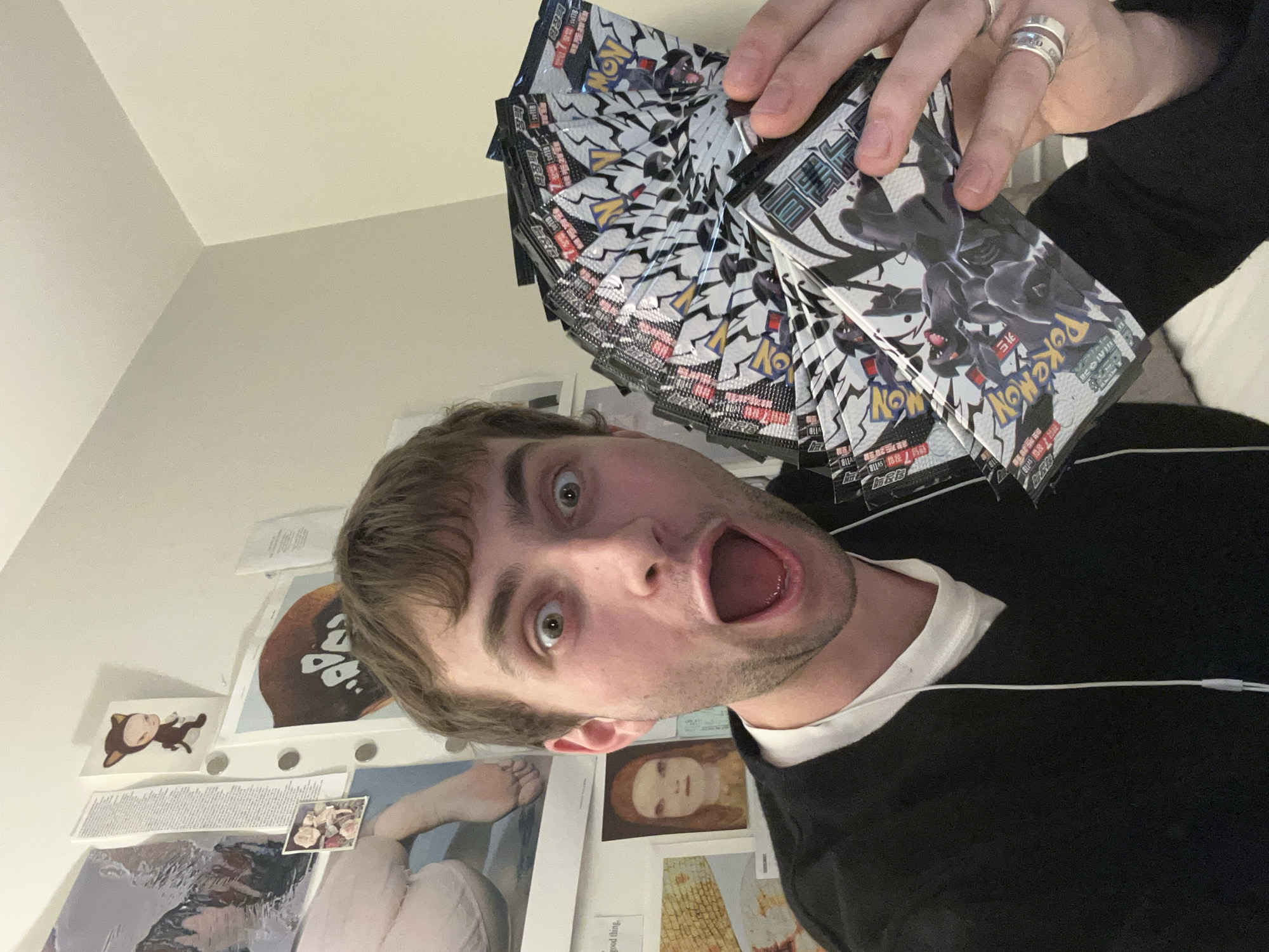 A selfie taken indoors, likely in a residential setting. A fair-skinned young man with short brown hair and wide eyes is holding a fan of unopened Pokémon trading card game booster packs in his right hand. He is wearing a black long-sleeved shirt with a white collar visible underneath and a white earbud cord is visible across his chest. His mouth is open in a surprised or excited expression. He has a few rings on his fingers.
The background is a white wall covered with various pictures and items affixed to it, including what appears to be artwork, photographs, and other printed materials. The room appears to be well-lit, possibly by artificial light. There's no readily identifiable landmark or element indicating the location is London. Therefore, it's not possible to identify the specific location as London, United Kingdom based solely on the provided image.