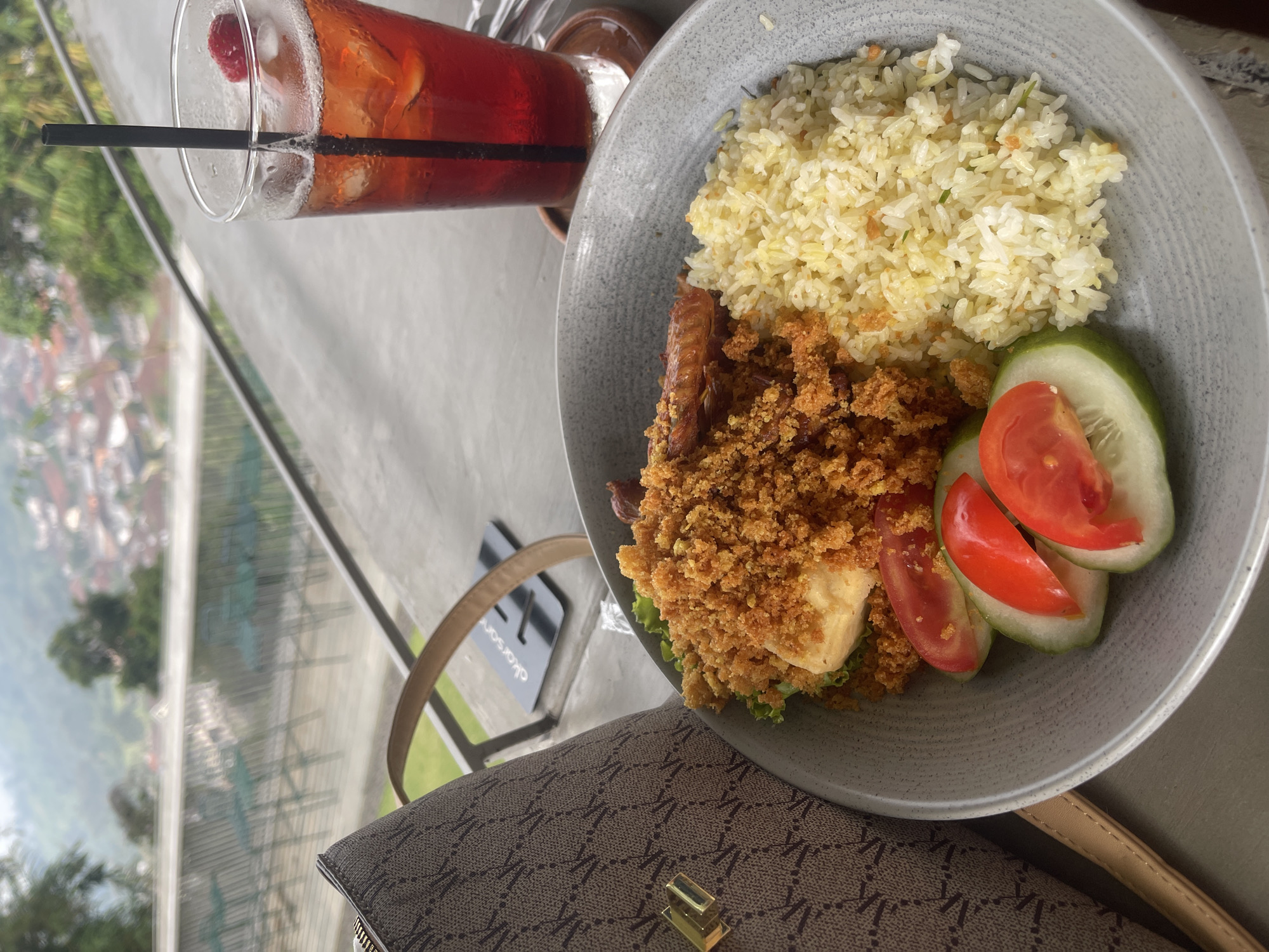An overhead medium shot captures a meal served on a textured gray plate. The meal consists of fried rice, a piece of fried chicken or tofu covered in crispy crumbs, and garnishes of sliced tomatoes and cucumber. To the right of the plate is a tall glass filled with a red beverage, ice cubes, and a raspberry. A black straw is submerged in the drink. The food and drink are placed on a concrete surface, possibly a table, next to a brown patterned bag. In the background, a scenic view of a village nestled in a valley with lush green hills and trees unfolds. The setting appears to be an outdoor dining area, offering a panoramic vista. The location is confirmed as Pecalukan, Indonesia, based on the provided context.