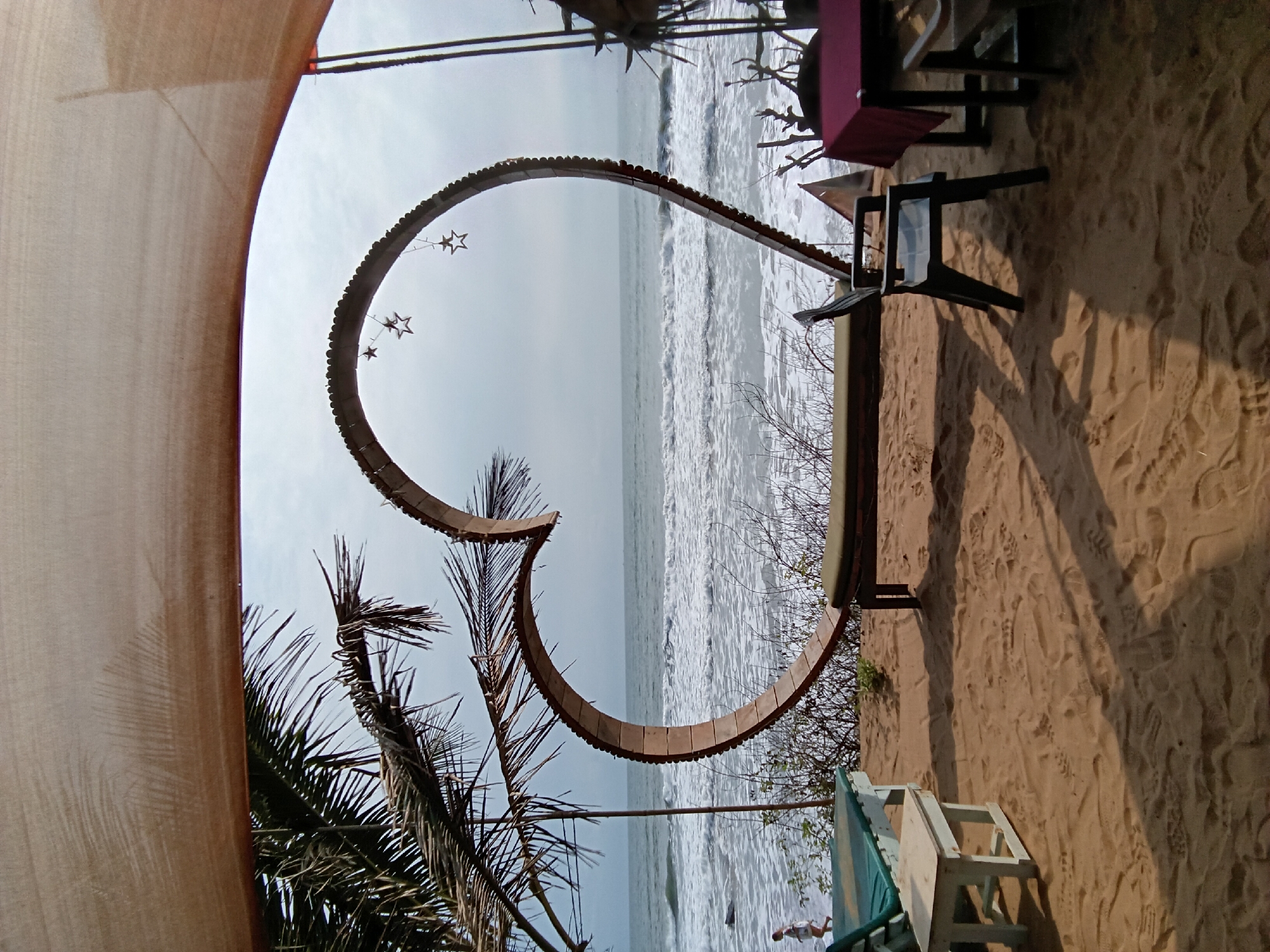 The image presents a scene at Ashvem Beach in Goa, India. The view is partly framed by a beige canopy or curtain in the upper left. A large, heart-shaped structure made of dark-brown wood segments is the focal point, adorned with dangling star-shaped ornaments. The sea is visible through the heart, with waves rolling onto the shore. Palm trees are present on the left side, adding to the beach environment.
The right side of the image shows a sandy beach area with footprints. Dining furniture, including chairs and a table with a maroon tablecloth, is situated on the sand. Additionally, a partially visible green and white beach chair rests on the sand. A sparse array of dry branches and vegetation can be seen near the base of the heart-shaped structure. There is a human figure in the ocean as well.
