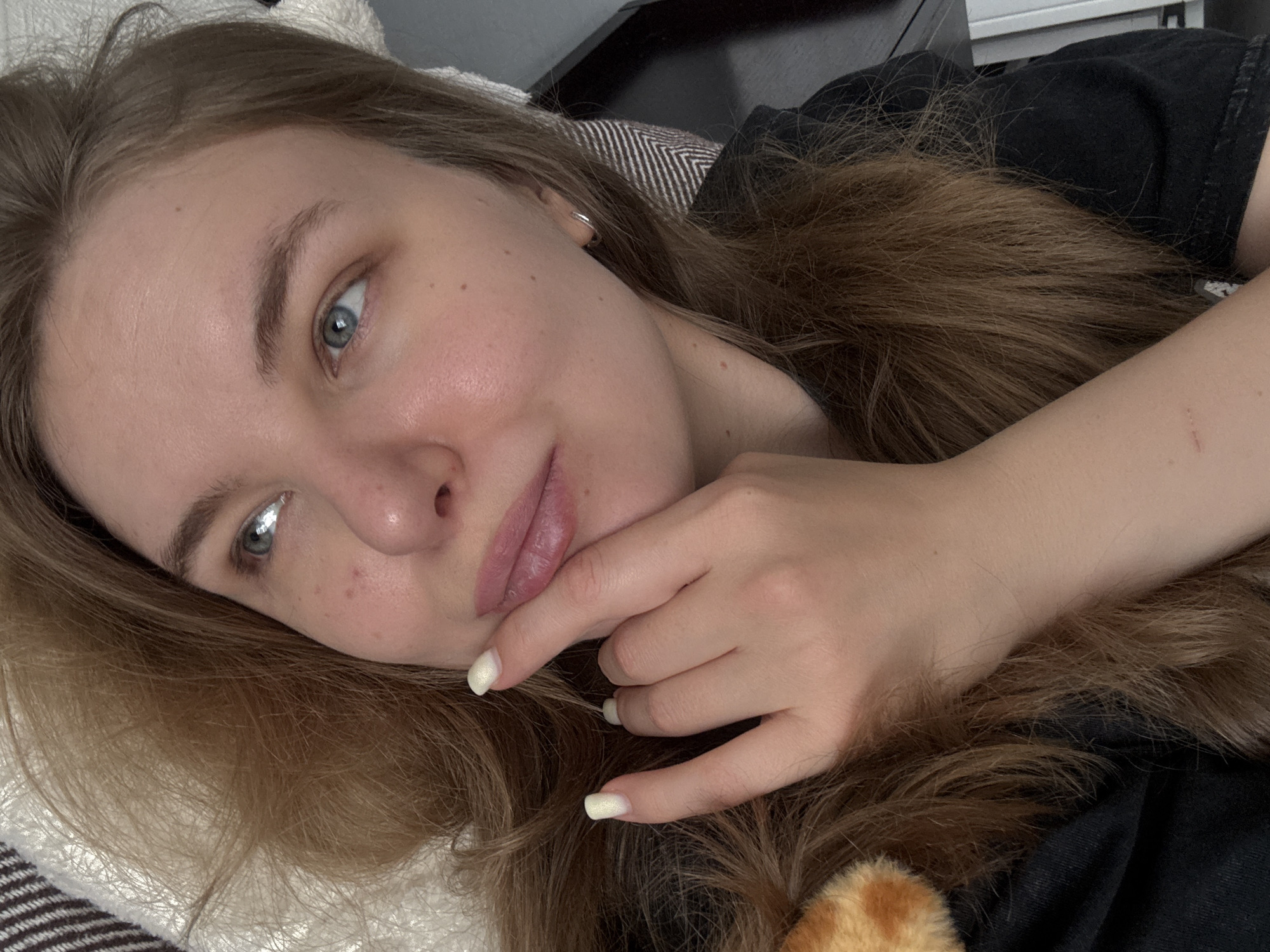 A close-up, tilted selfie depicts a young woman with long, light brown hair, fair skin, and blue eyes. She is lying down, with her head resting on a white, textured pillow. Her right hand is raised, with her index finger touching her lower lip. She wears a dark-colored top. Part of a black object is in the background. There is no evidence indicating the location.