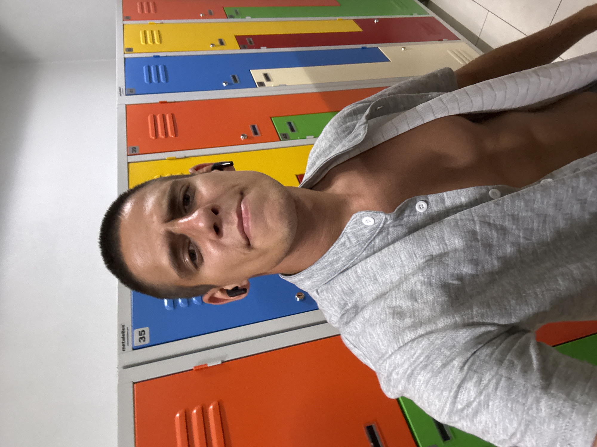 The image is a selfie taken in a locker room. The subject is a man with a buzz cut, wearing an unbuttoned grey shirt, revealing his chest. He has black wireless earbuds in his ears. Behind him are rows of brightly colored lockers, predominantly orange, blue, yellow, and green. The lockers are labeled with numbers such as 35, 39, and 40. The floor appears to be tiled. The overall setting suggests a gym or a similar facility. The image does not contain any identifiable landmarks or clues to place it specifically within Sofia, Bulgaria.