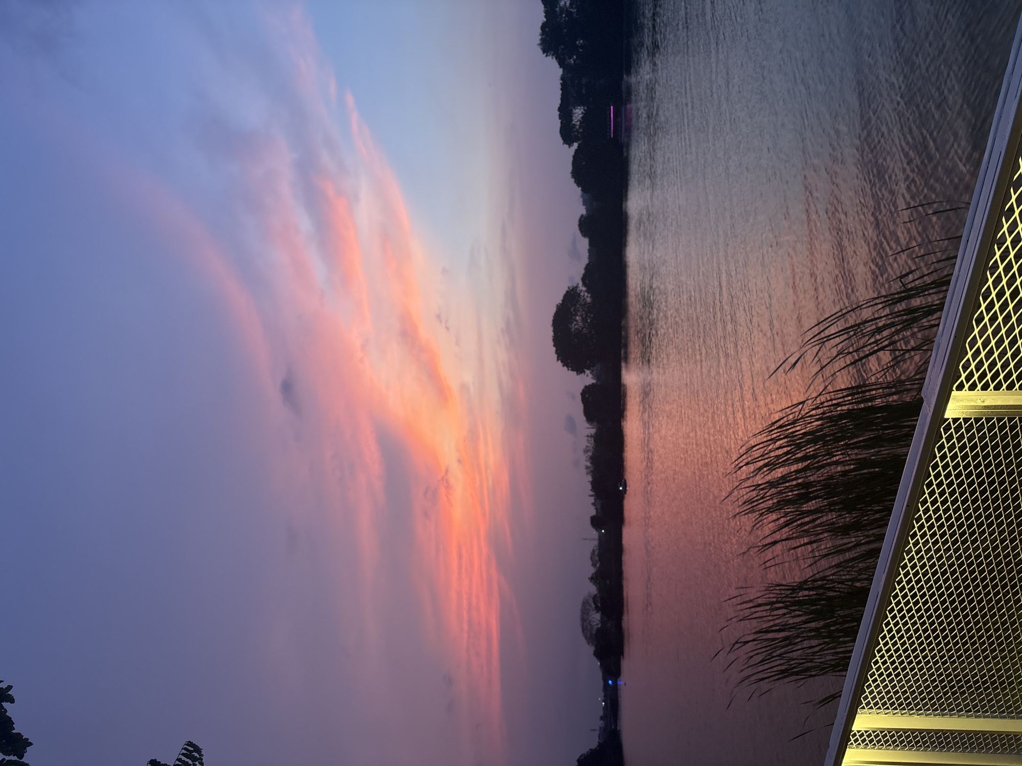 The image depicts a sunset scene at a body of water, likely a lake or reservoir, in West Jakarta, Indonesia. The sky displays vibrant colors, with streaks of orange and pink blending into the blue and purple hues of dusk. The water reflects the colors of the sky, creating a mirrored effect. A line of trees and vegetation is visible along the opposite shore, with subtle lighting suggesting urban structures in the distance. In the foreground, architectural elements are present, possibly a structure or building with a lattice-like design and vegetation obscuring a portion of the frame. The orientation of the image suggests it was taken vertically.