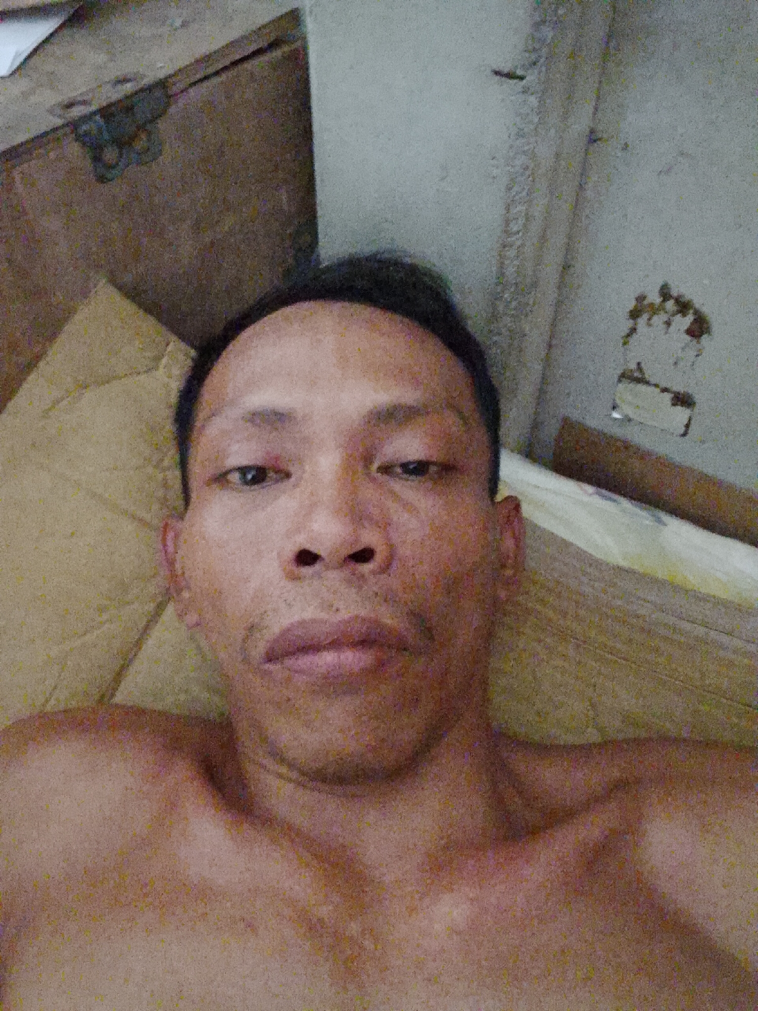 A bare-chested person with short black hair and dark eyes is looking directly at the camera. The person's skin tone appears to be tan. They are lying on a yellowish or tan textured surface, possibly a bed or cushion. Above them, there is a brown wooden surface with a metallic clasp and a dirty, white wall with discoloration and debris. The location is Bukit Ketok, Indonesia. The scene suggests an indoor setting, possibly a bedroom. There are no visible actions or interactions other than the person posing for the camera.