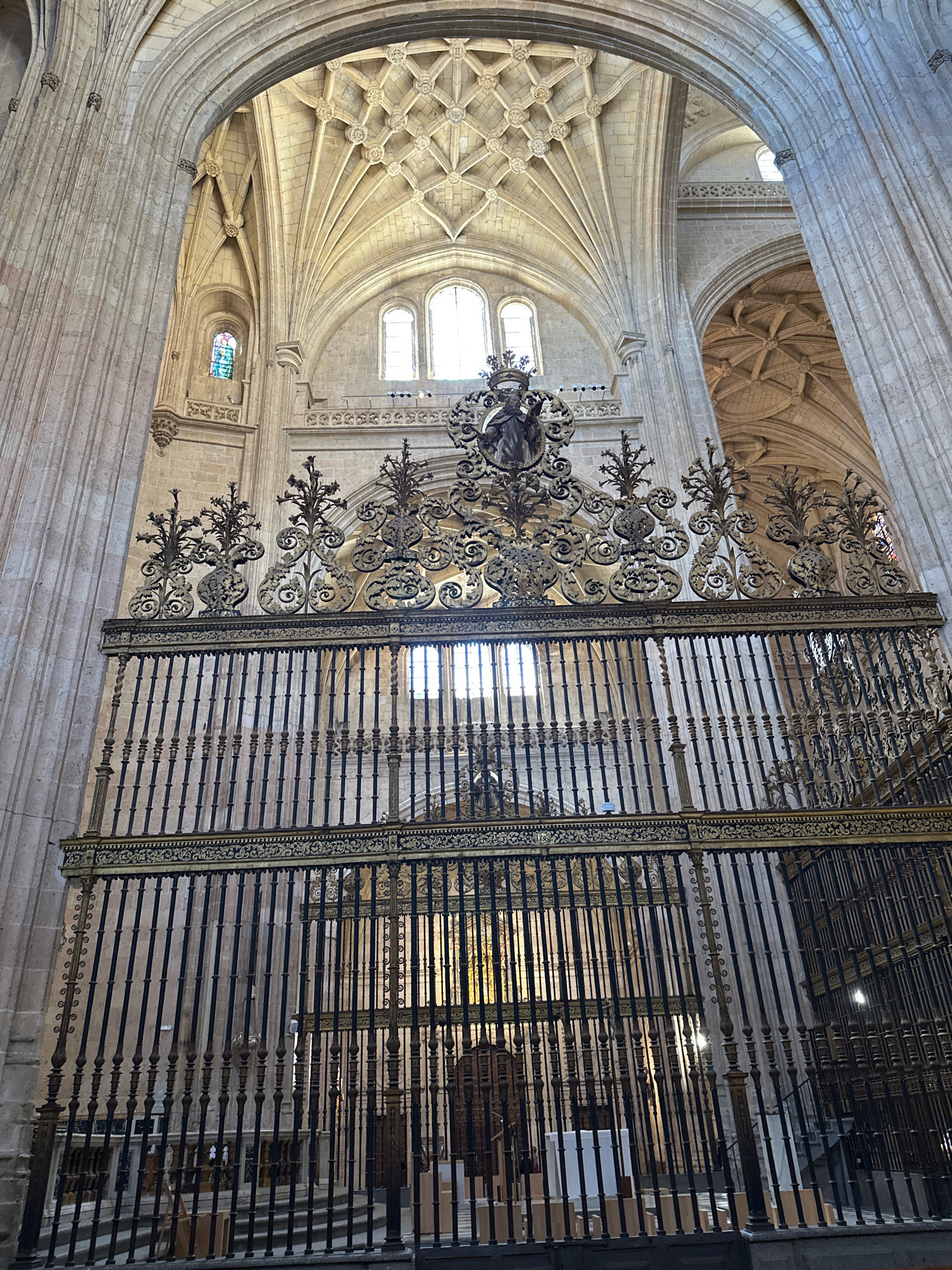 An interior view of a cathedral in La Lastrilla, Spain, showcases intricate Gothic architecture and ornate metalwork. The image is dominated by a large, black wrought-iron screen adorned with gilded decorative elements and a central sculptural figure wearing a crown. Behind the screen, a dimly lit nave with marble flooring and wooden pews is visible, suggesting a place of worship. The vaulted ceilings, characteristic of Gothic cathedrals, are a prominent feature, with ribbed vaults and ornamental plasterwork. Sunlight streams through high windows, illuminating stained-glass panels in the distance. The overall impression is one of grandeur and historical significance.