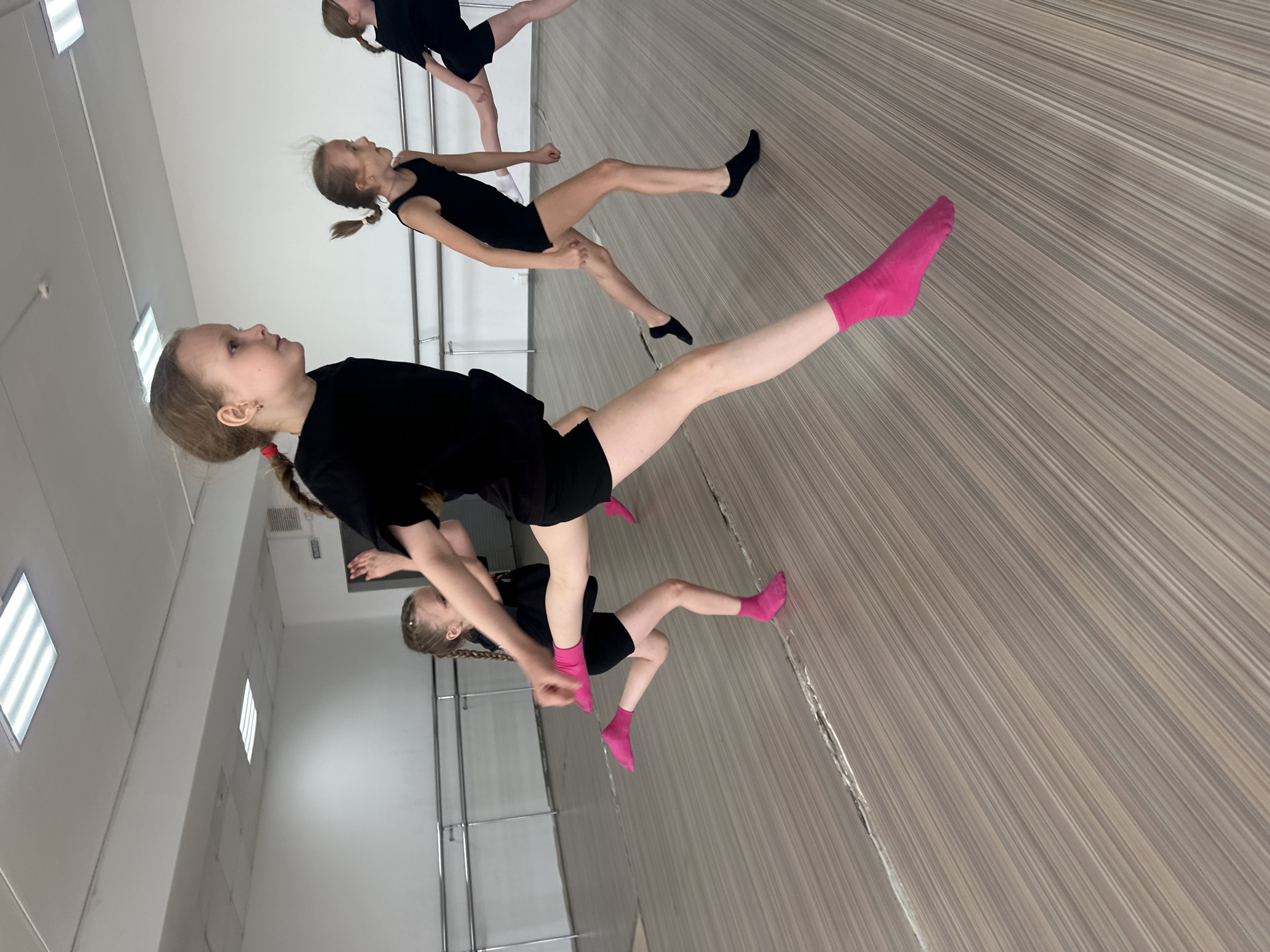 The image depicts a dance studio with multiple young girls practicing ballet. They are all wearing black leotards and some have pink socks on their feet. The studio has a light-colored floor with a striped pattern and a white wall with ballet barres. The location is presumed to be in Votkinsk, Russia.