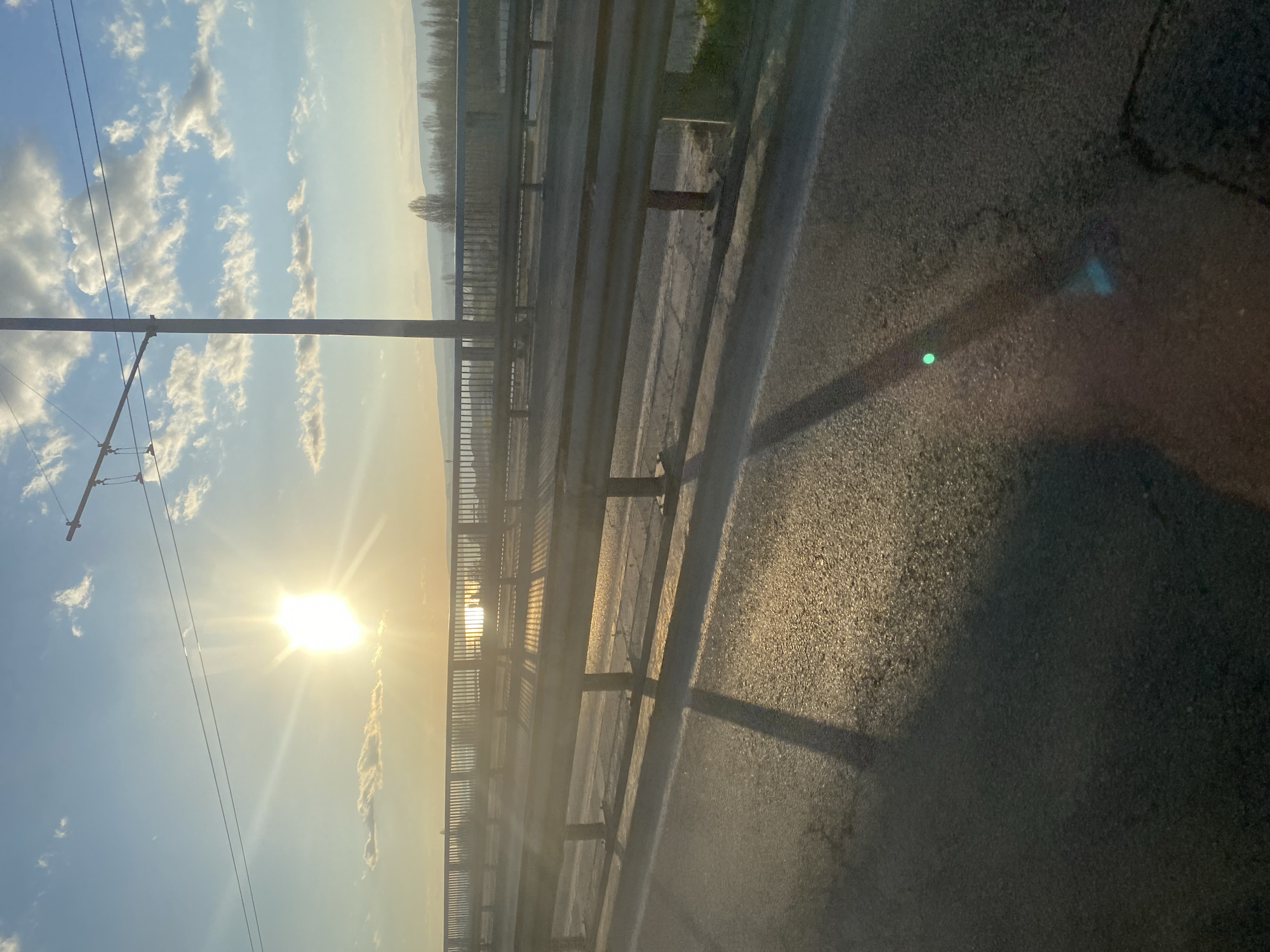 The image depicts an outdoor scene at either sunrise or sunset, characterized by the sun's low position and golden light. The sky is a clear blue with scattered cumulus clouds, especially prominent on the left. The sun, intensely bright and causing significant glare and lens flare, dominates the upper right portion, casting long shadows and bright reflections.
In the middle distance, a gentle range of hills or mountains stretches across the horizon, with a solitary tree visible against the lighter sky, slightly to the left of the image center. On the far left, a utility pole with multiple overhead wires extends vertically, suggesting the presence of public transport infrastructure.
The central and right parts of the image feature a section of what appears to be an elevated road or bridge. A sturdy metal guardrail, composed of horizontal beams supported by vertical posts, runs along its edge. The asphalt road surface, showing texture and some cracks, is visible in the foreground and beneath the guardrail. The sun's light intensely illuminates parts of the road and the top surfaces of the guardrail, while other sections are in deep shadow. Distinct green and blue light artifacts are visible on the shadowed asphalt in the immediate foreground, likely lens flares or internal reflections from the vehicle the photo was taken from. The scene conveys a sense of travel through a scenic area, consistent with the landscape near Varna, Bulgaria. No text is visible.