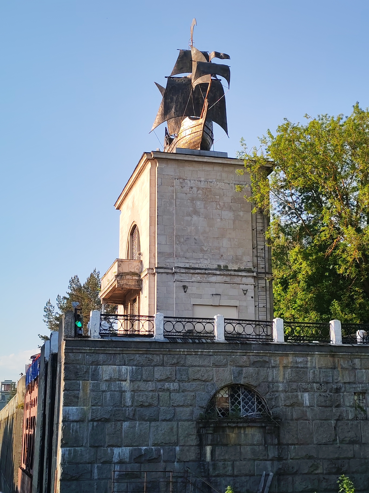 The image shows a stone structure topped with a metal sculpture of a ship, located in Yakhroma, Russia. The structure is multi-tiered, with a base constructed of large gray stone blocks featuring an arched window covered with decorative metal grilles. Above this, a lighter-colored stone building with a balcony is situated, and a metal ladder is attached to its side. A black metal railing with white posts surrounds the upper platform. A green traffic light is visible next to the railing. The sculpture of the ship sits atop the building, its black sails spread against the clear blue sky. Green trees are seen to the right of the structure. There are no people visible.