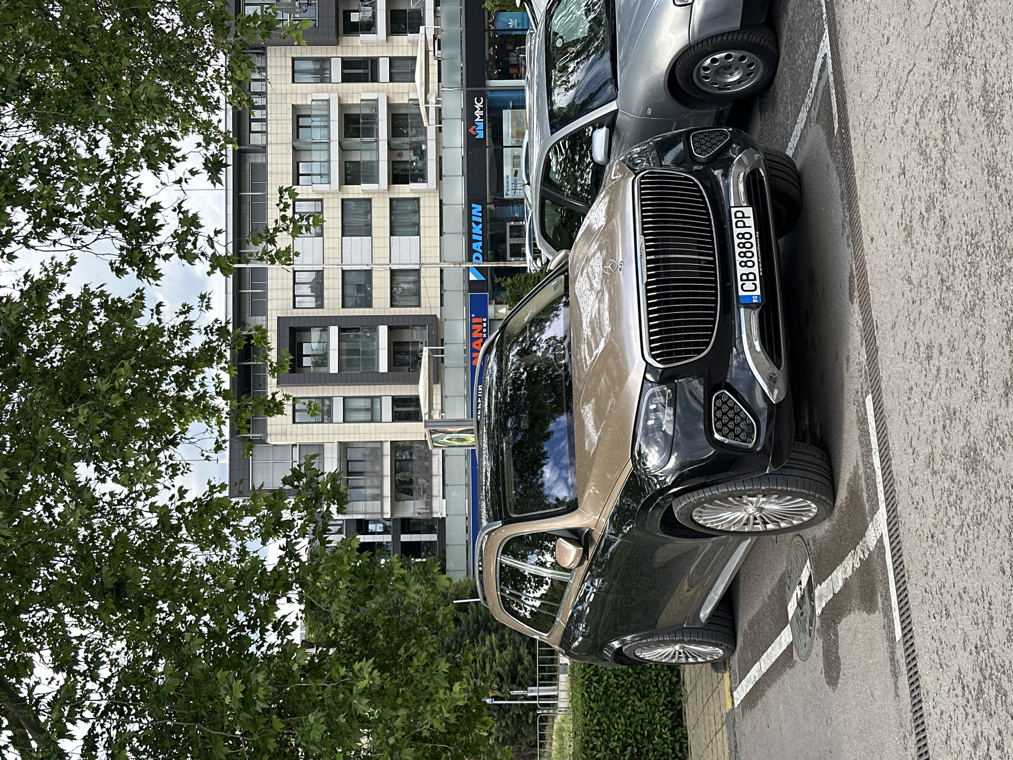 The image depicts a black and gold-colored Rolls Royce Cullinan SUV parked in a designated parking space next to a silver-colored vehicle. The license plate of the Cullinan reads "CB 8888 PP" and is Bulgarian. The backdrop consists of a multi-story building with a modern facade featuring a combination of white and gray panels, and numerous windows. Signage visible on the building includes "VM MMC" and "DAIKIN", indicating commercial establishments. The foliage of a green tree partially obscures the left side of the frame. The location appears to be an urban area in Varna, Bulgaria, as indicated by the license plate number. The weather conditions appear to be overcast with diffuse lighting. There are no people in the immediate view.