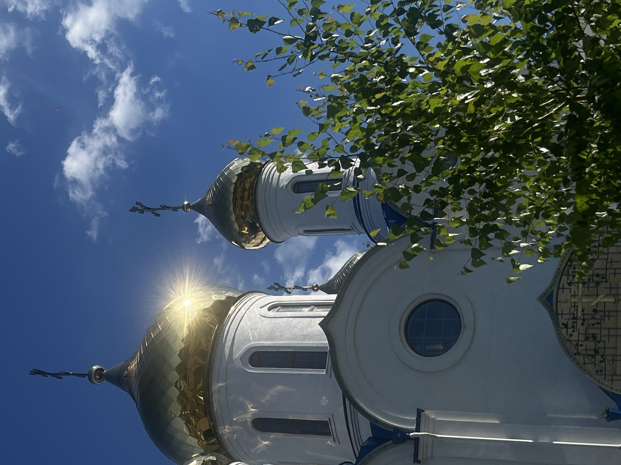 A low-angle, eye-level shot captures the sun-drenched facade of a white Orthodox church with golden onion domes and crosses, situated in Novopokrovskaya, Russia. The largest dome, positioned on the left, reflects intense sunlight, creating a prominent starburst flare. A smaller golden dome with a cross is visible to its right. The church features tall, arched windows on its upper sections and a circular window lower down on the main structure. Blue accents are visible beneath the domes and along the roofline. A green-leafed tree partially obscures the right side of the church and the smaller dome in the foreground. The sky is clear blue with scattered white cumulus clouds. A small, magenta-colored, elongated object is visible high in the sky, between the two domes. No people are present.