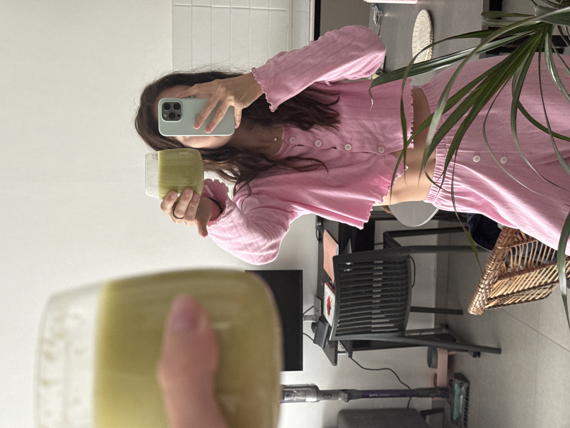 A person in pink loungewear is taking a selfie in a room. They are holding a glass of green liquid. The background includes a white tiled wall, a desk with a computer monitor, a chair, a plant, and a vacuum cleaner. The location is not discernible from the image.