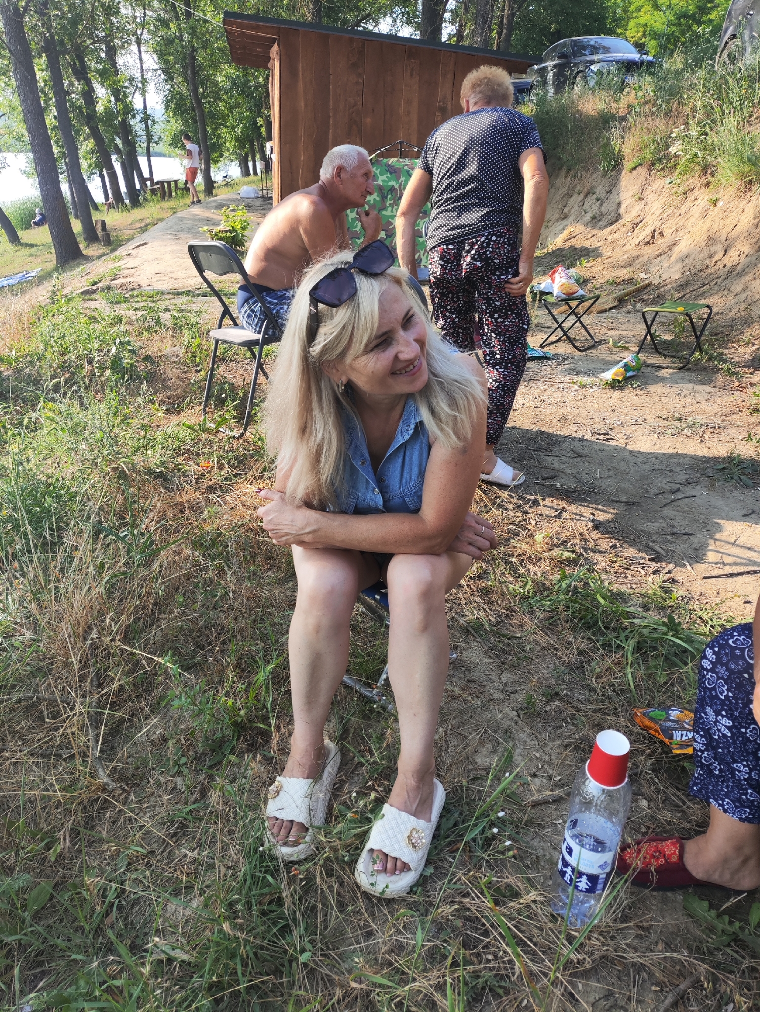 The image depicts an outdoor scene with several individuals and objects on a grassy, sandy embankment, likely near a body of water, consistent with a location in Găleștii Noi, Moldova. In the foreground, a woman with blonde hair and sunglasses on her head is seated, wearing a denim shirt and sandals. She is looking to her right and smiling. Behind her, two other individuals are visible. One, a man with white hair, is seated on a folding chair, shirtless and wearing checkered shorts, looking to his left. Another person, wearing a black polka-dot shirt and patterned pants, stands facing away from the camera.
Scattered around the area are several objects, including folding chairs, a wooden structure resembling a small shed or cabin, a plastic bottle with a red cup on top, bags of chips, and other miscellaneous items. The environment is characterized by dry grass, trees, and a dirt path. The overall impression is of a casual gathering or picnic in a natural setting.