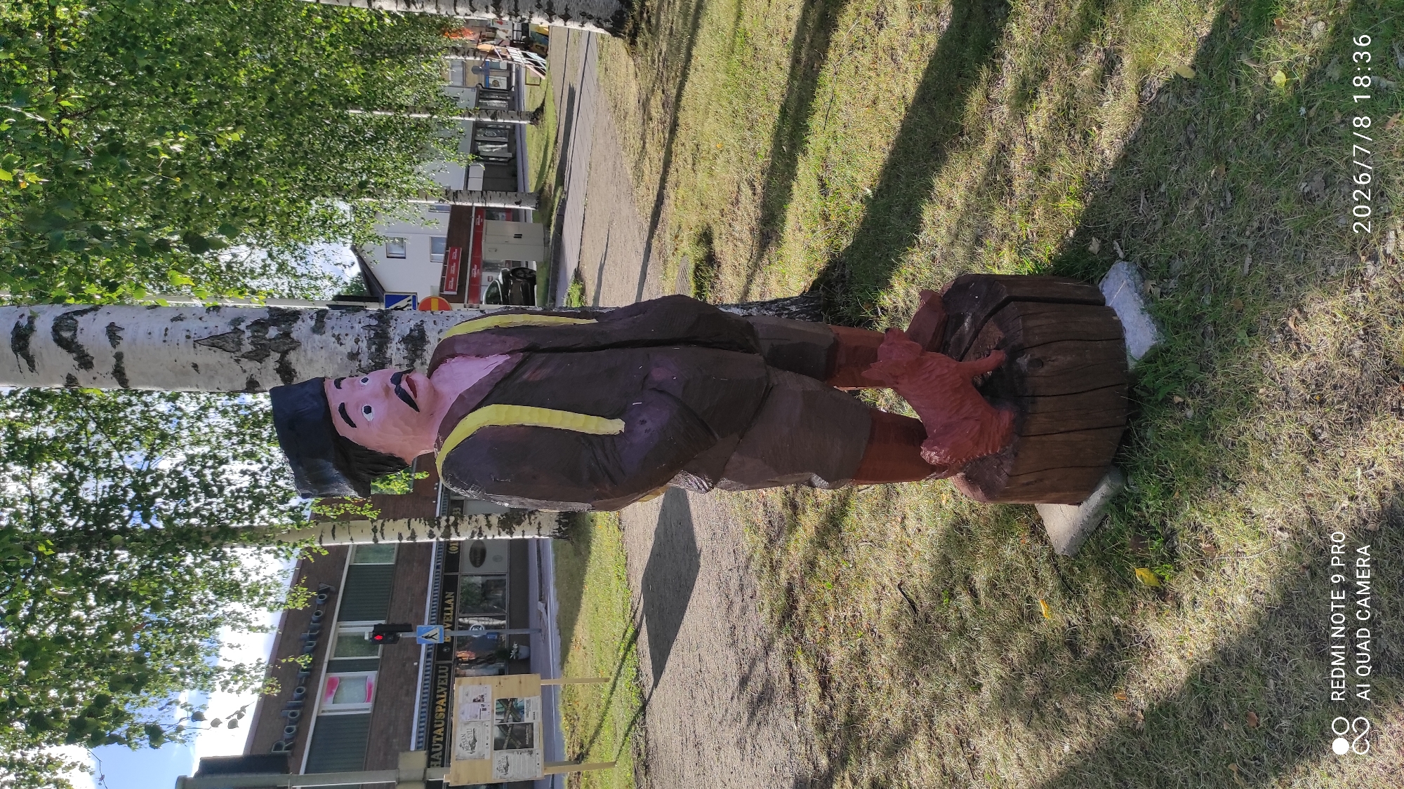 A wooden folk art statue of a man with a dark hat, brown jacket, and shorts, stands on a wooden log base in front of a birch tree. A small red dog sculpture is at the base of the log. The statue is located on a sidewalk in Iisalmi, Finland, with a street and buildings in the background. The buildings include shops with signs in Finnish, such as "Radio-outro Oy" and "Hautauspalvelu". The sky is blue and sunny with trees providing shade. The bottom of the image shows a watermark with "REDMI NOTE 9 PRO AI QUAD CAMERA" and a date "2026/7/8 18:36".