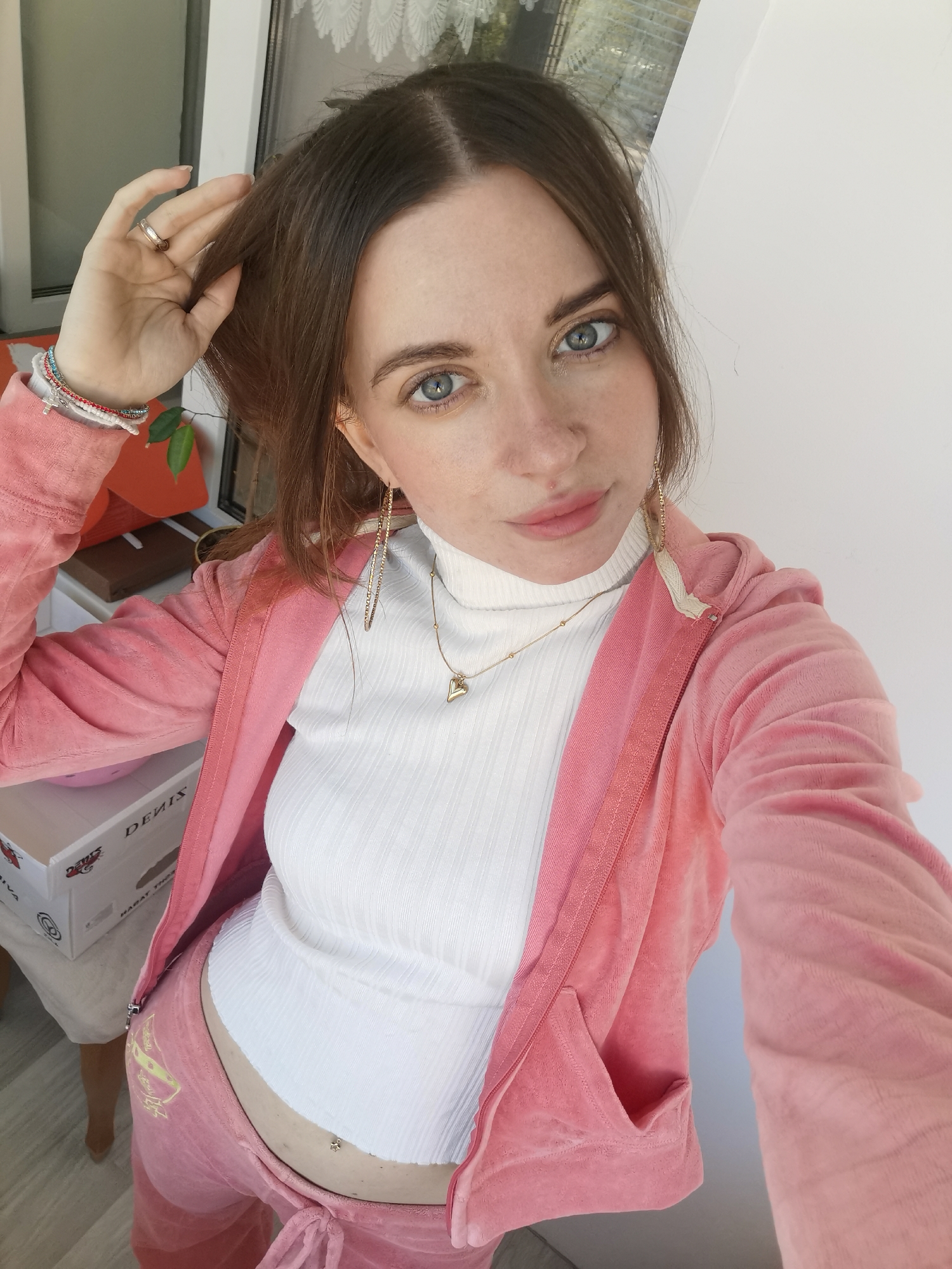A selfie shot depicts a woman indoors. She has light skin, blue eyes, and brown hair. Her left hand is raised, touching her hair. She wears a white turtleneck shirt, a pink velour tracksuit jacket and pants, gold hoop earrings, and a gold heart necklace. A gold ring is on her ring finger, and bracelets are on her left wrist. A gold star-shaped piercing is visible on her stomach. A "DENIS" branded box is behind her. A window with white curtains is visible in the background. The location is Chaikovsky, Russia.
