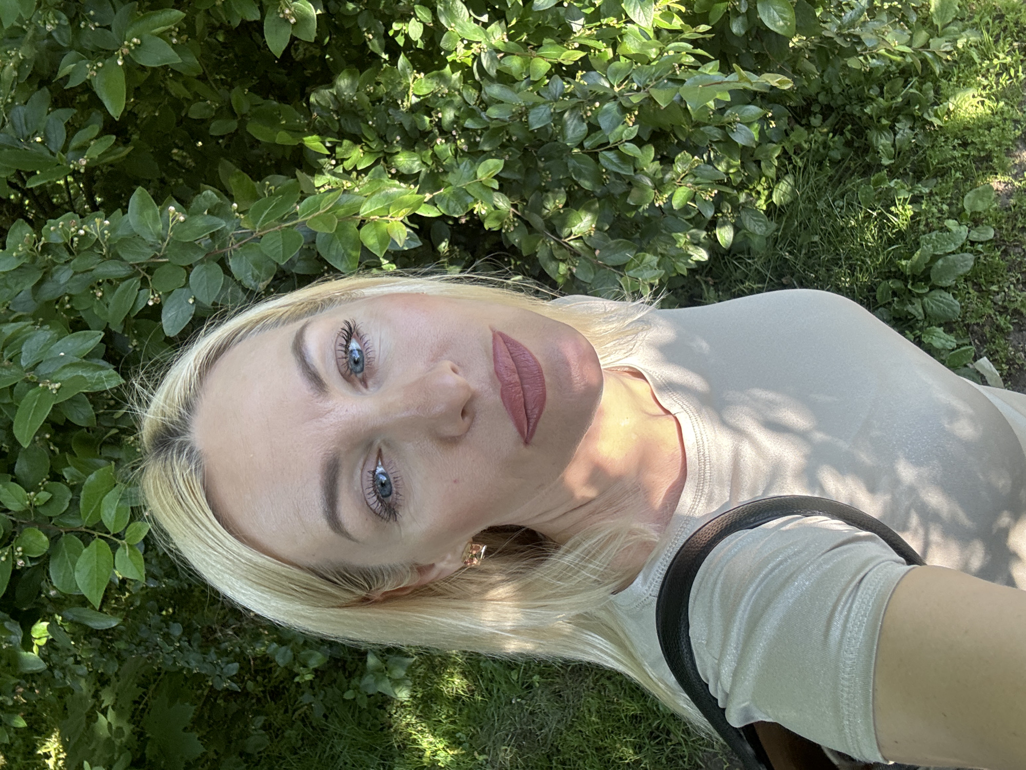 A close-up outdoor portrait features a blonde woman with blue eyes, looking directly at the camera. Her head is angled, and she appears to be lying down or leaning back, with lush green foliage and grass forming the background. She has long, dark eyelashes, and her lips are adorned with a matte pinkish-brown lipstick. Her blonde hair frames her face, with visible roots. She is wearing a light-colored, possibly shimmery, short-sleeved top, and a black strap, likely from a bag, is seen across her right shoulder and arm. A small golden earring is visible on her left ear.
The setting is a verdant outdoor area, abundant with green leaves and bushes, some featuring tiny white buds, and patches of grass. The scene is brightly lit by natural sunlight, casting distinct shadows and highlighting parts of the foliage and her attire, suggesting a clear day, likely during the mid-day or early afternoon. Her expression is neutral and direct. No visible text is present in the image.