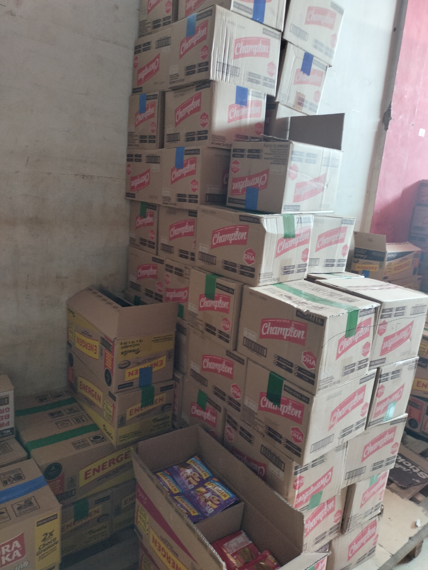 The image shows a large stack of cardboard boxes, predominantly displaying the brand "Champion" in red lettering, some with "DHA" also visible. Several boxes are also branded "ENERGEN". The boxes are piled high in a corner with a light-colored, textured wall on the left and a pinkish-red wall on the right. Some boxes appear to be open, revealing packaging or contents. The scene is indoors, likely a storage area, possibly a warehouse or a shop backroom. The presence of multiple stacks of branded goods suggests a commercial or retail context. The overall impression is one of inventory and storage of packaged food or beverage products. The image does not contain any people. The lighting is somewhat dim, with shadows cast by the stacked boxes. The environment appears somewhat unkempt, with some boxes showing signs of wear. The location within Langsa, Indonesia is not identifiable from the visual information provided.